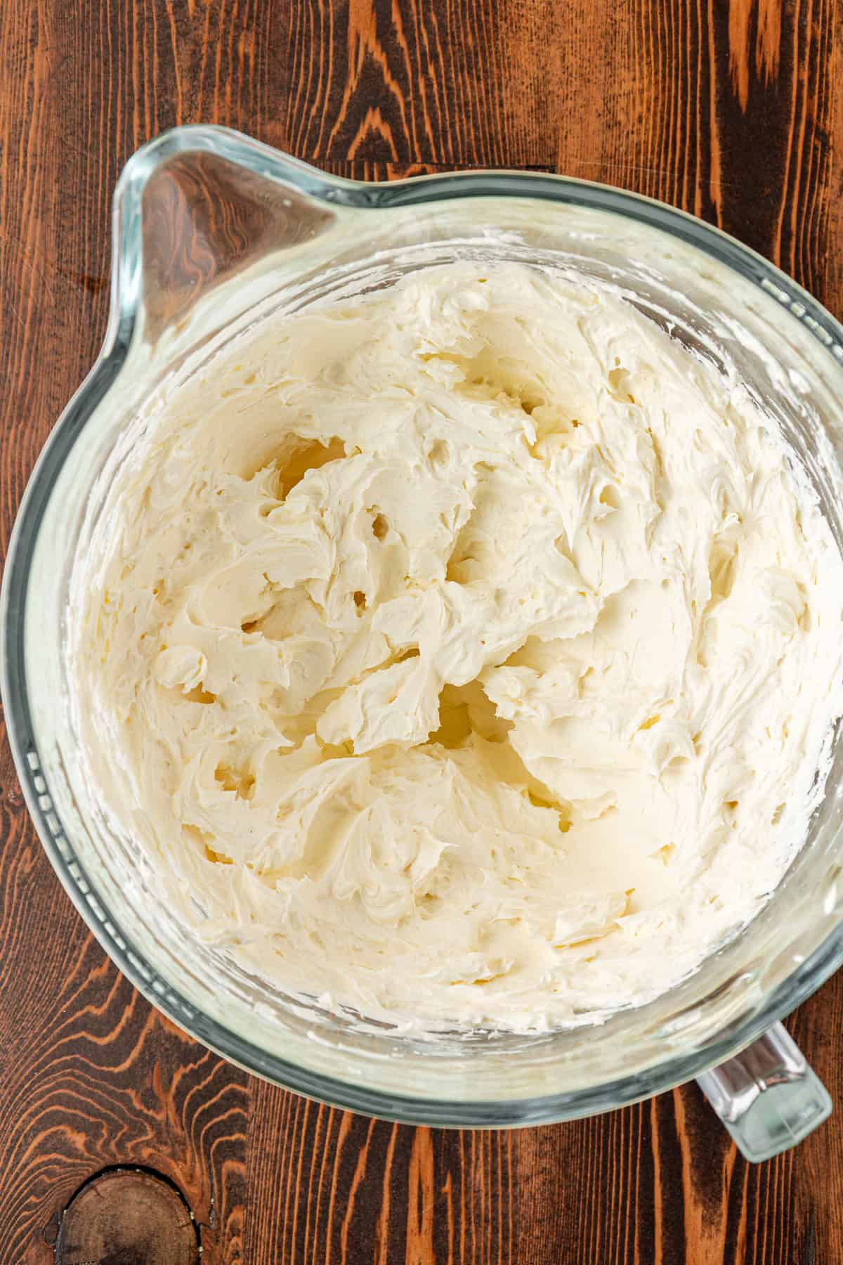 A glass mixing bowl filled with fluffy, whipped cream cheese frosting sits on a wooden surface. The frosting has a smooth and airy texture with visible peaks and swirls.