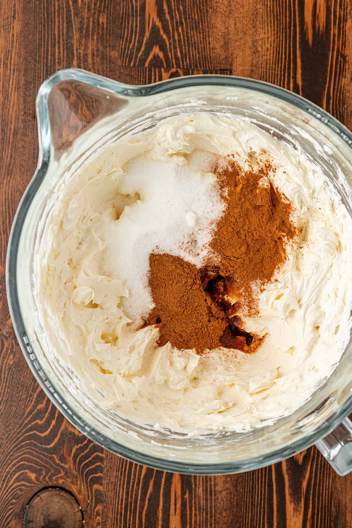 A glass mixing bowl on a wooden surface contains whipped cream cheese topped with granulated sugar, ground cinnamon, and vanilla extract, ready to be mixed together.