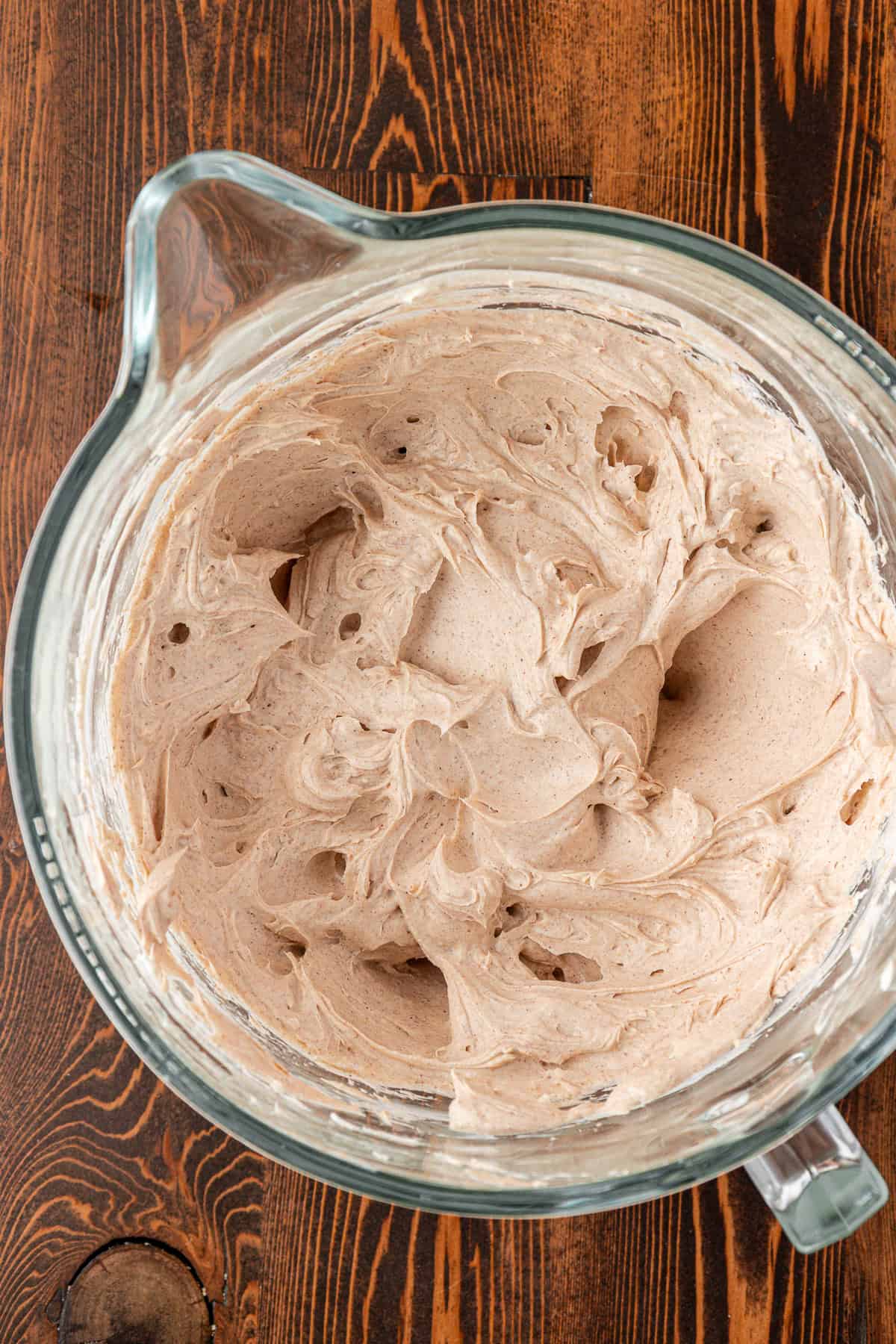 A glass mixing bowl filled with whipped, light brown frosting sits on a wooden surface. The frosting appears airy and smooth with soft peaks throughout.