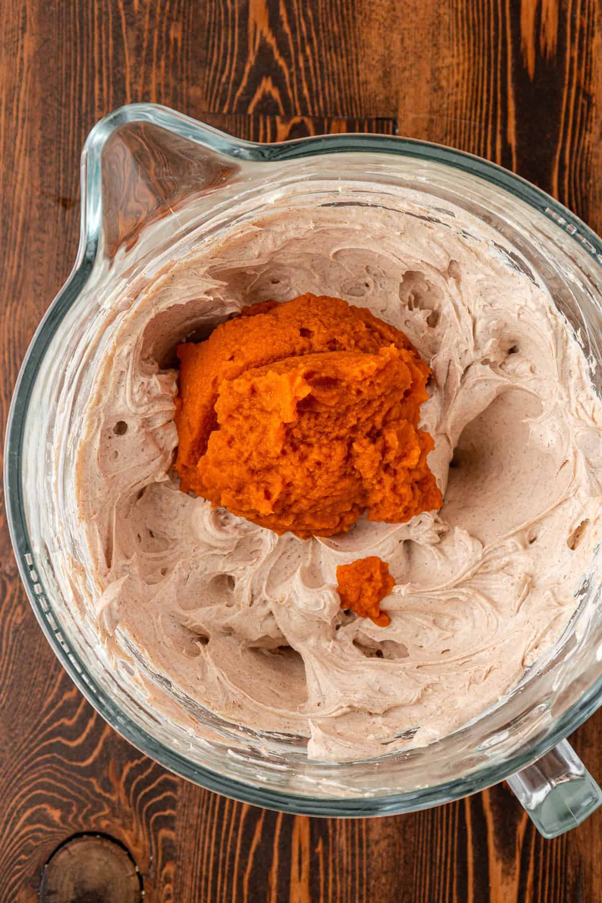 A glass mixing bowl on a wooden surface contains creamy whipped mixture with a mound of bright orange pumpkin puree on top.