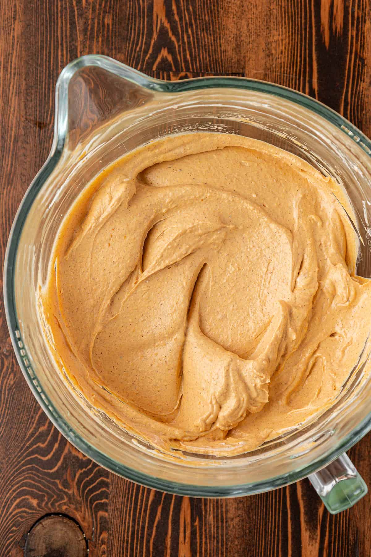 A glass mixing bowl filled with creamy, light brown batter sits on a wooden surface. The batter is smoothly blended with swirls visible on top.