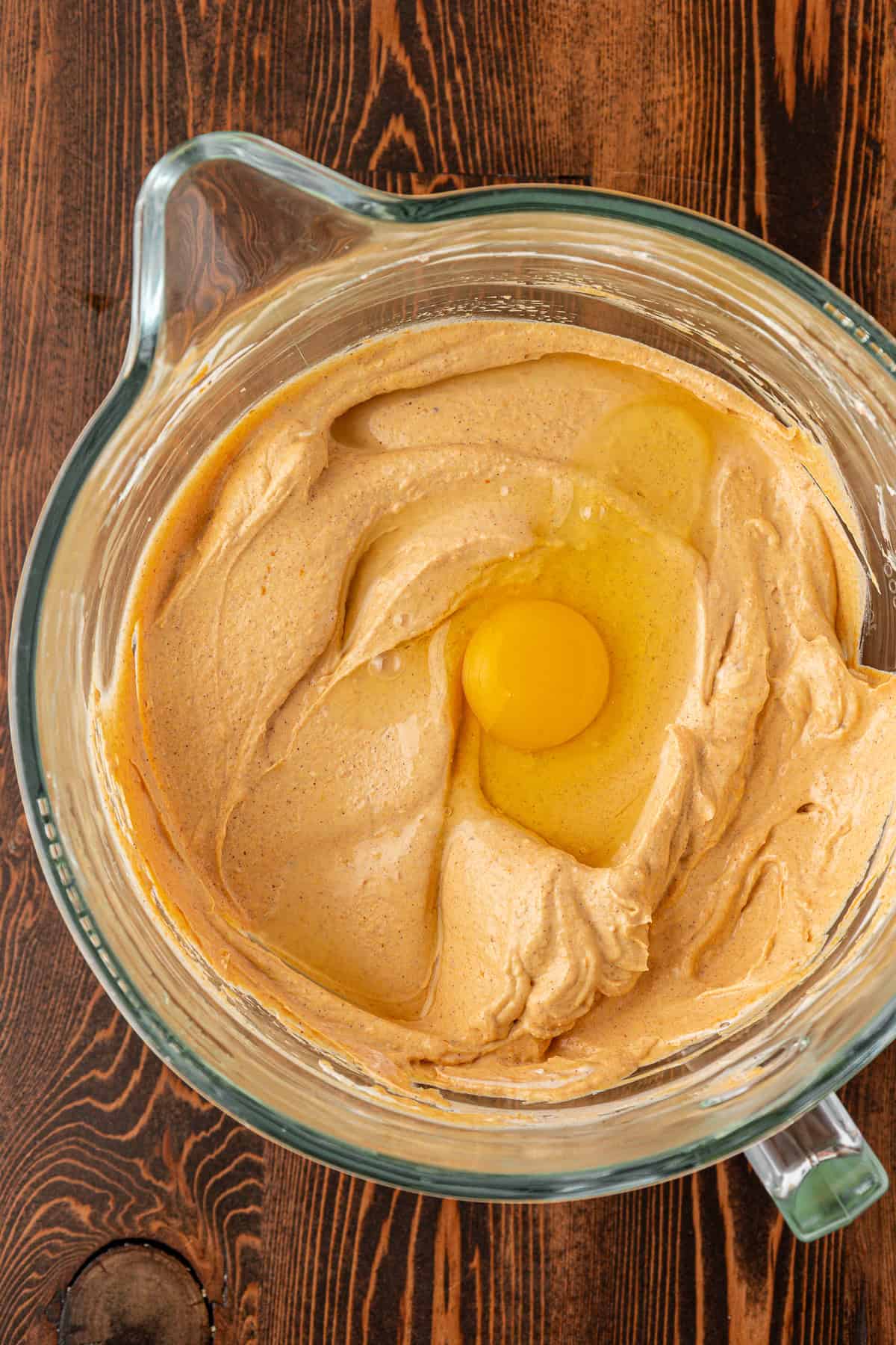 A glass mixing bowl with creamy light brown batter and a raw egg on top, set on a wooden surface.