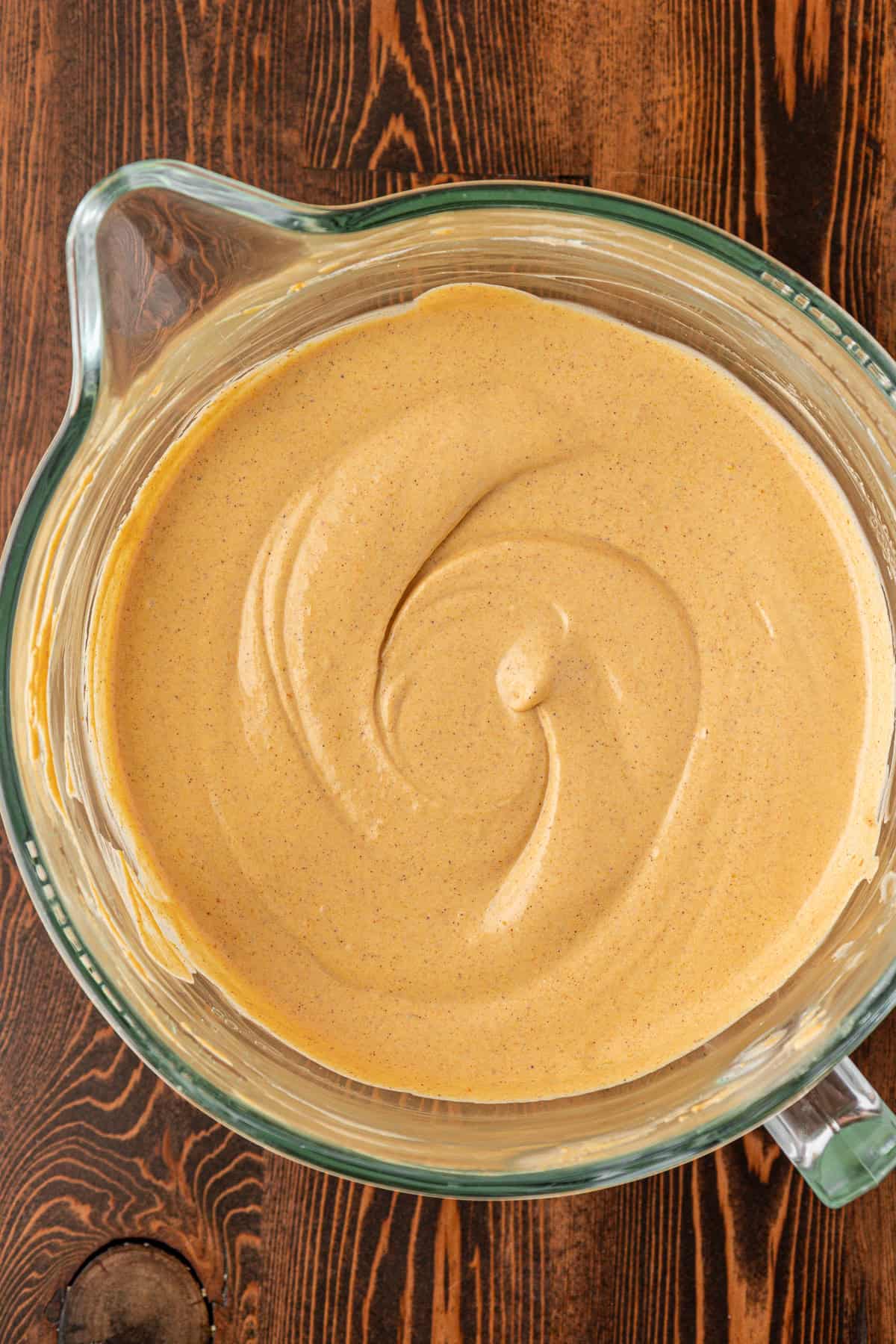 A glass mixing bowl filled with creamy, light brown pumpkin pie filling, swirled smoothly on top, sitting on a wooden surface.