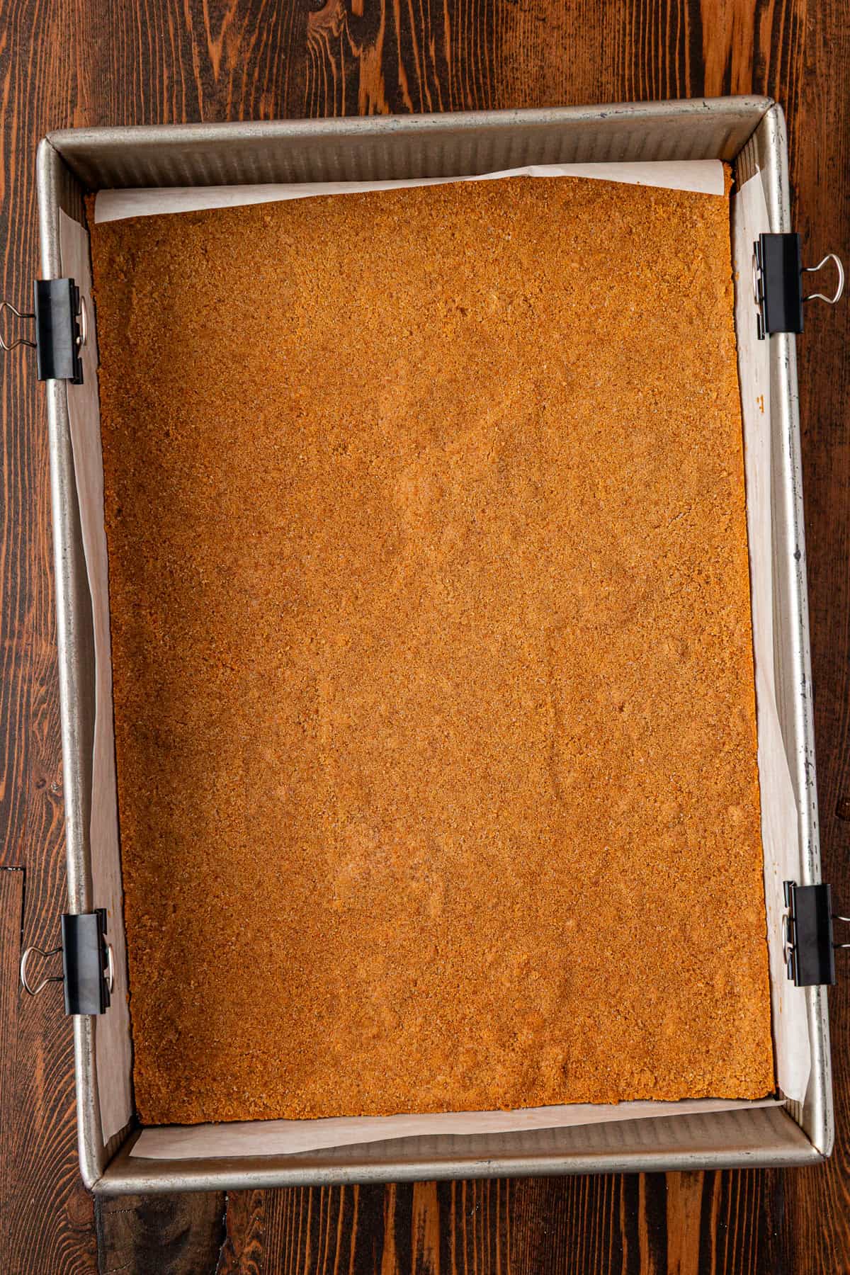 A square baking pan lined with parchment paper and binder clips holds a flat, golden-brown graham cracker crust on a wooden table.