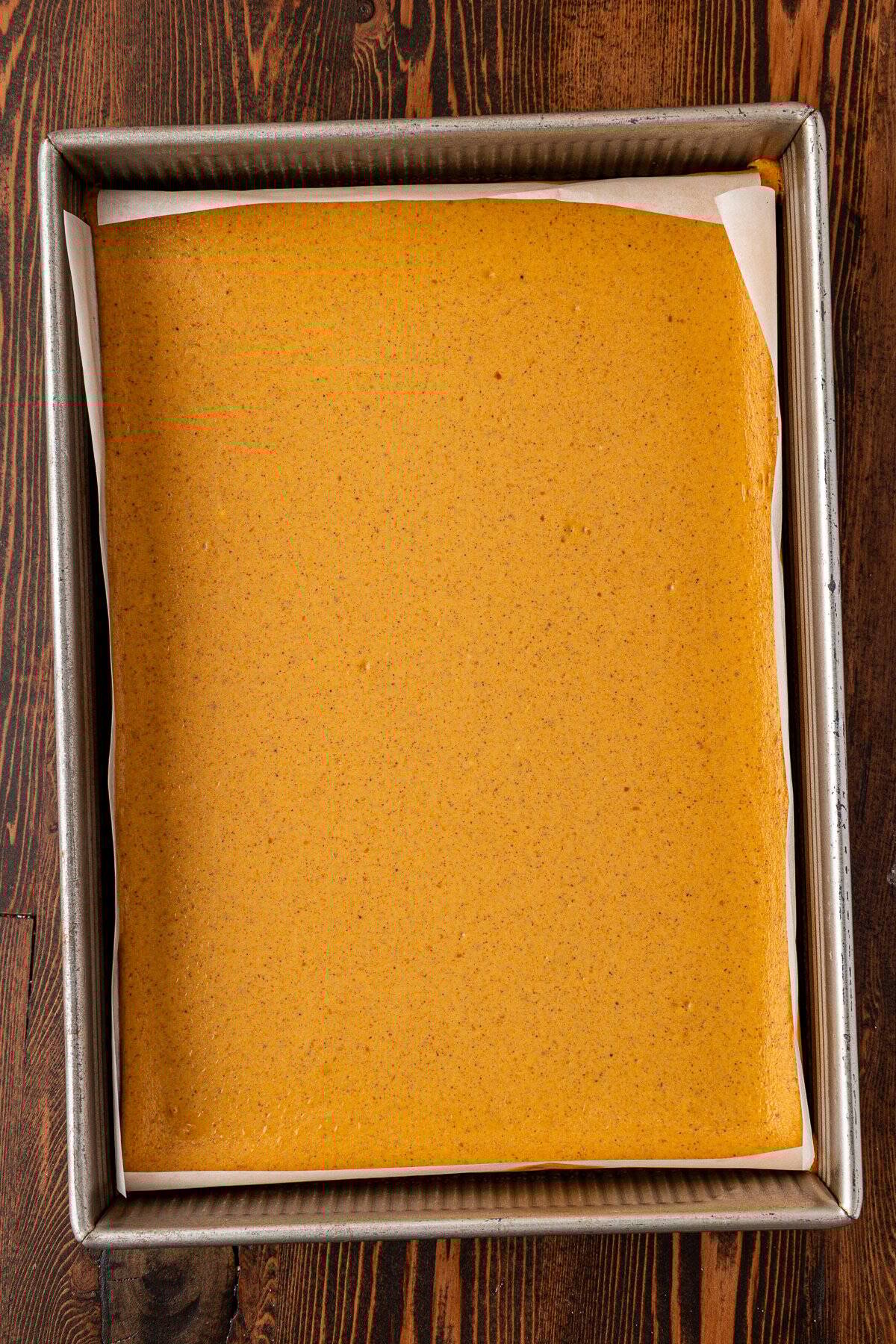 A rectangular baking pan lined with parchment paper, filled with a smooth, light brown batter, sits on a dark wooden surface.
