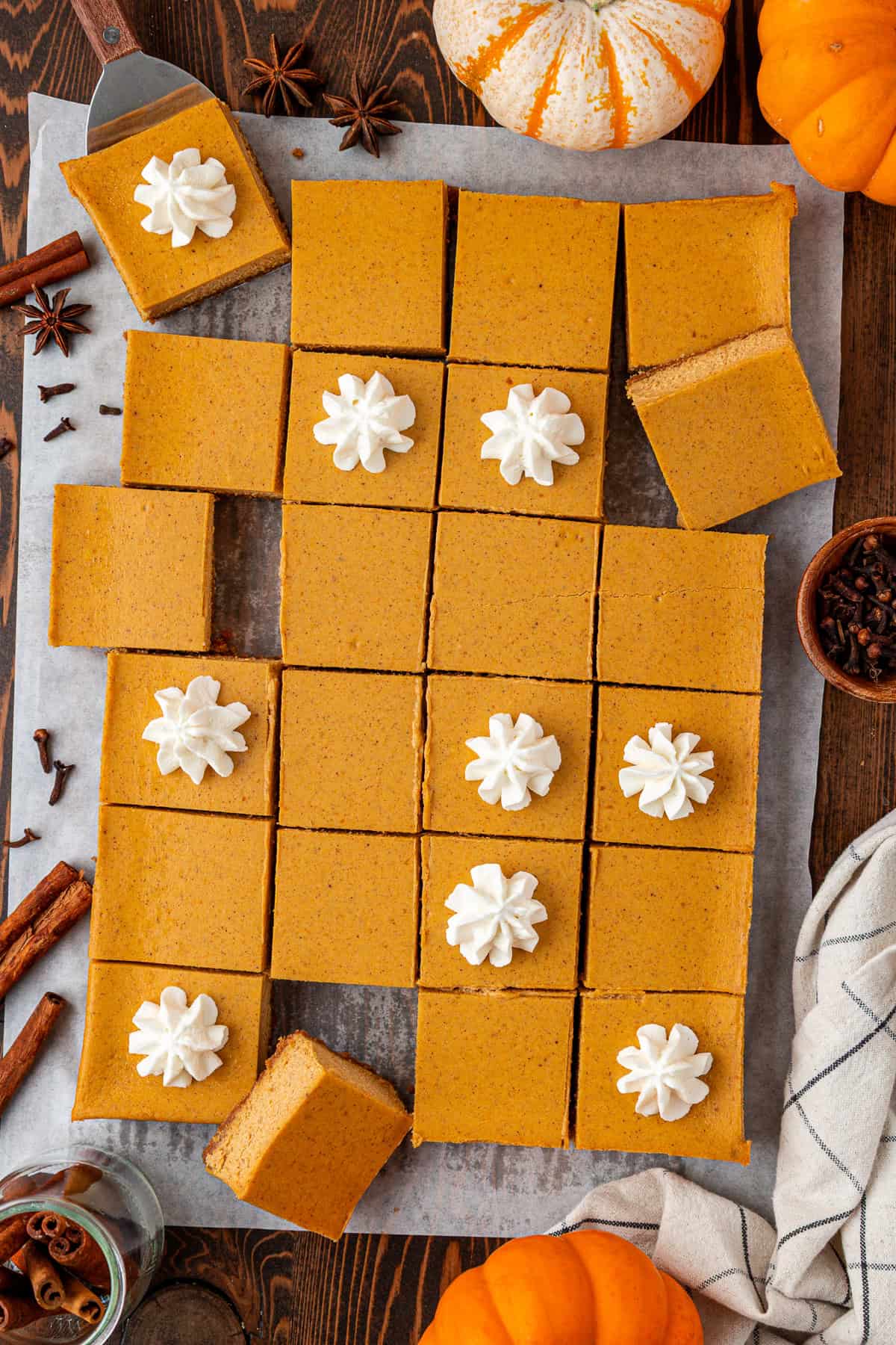 Top-down view of pumpkin bars cut into squares on parchment paper, some topped with whipped cream, surrounded by whole mini pumpkins, cinnamon sticks, star anise, and cloves on a wooden table.