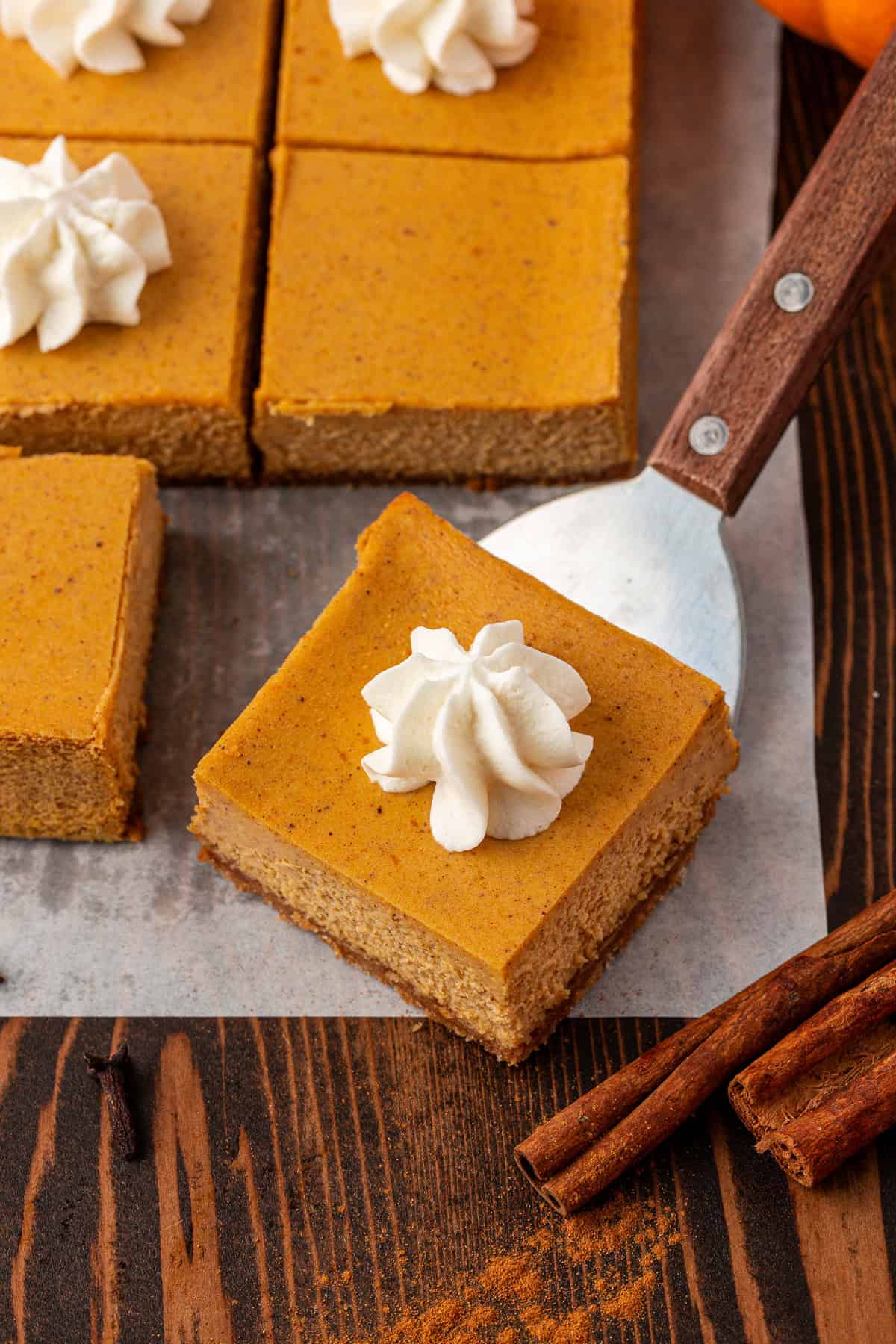 A close-up of pumpkin bars topped with whipped cream, arranged on parchment paper. One bar is being lifted with a spatula. Cinnamon sticks and a dusting of cinnamon are on the wooden table.