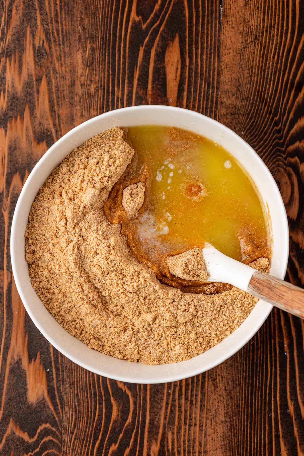 A white bowl filled with graham cracker crumbs and melted butter sits on a wooden table, with a spatula partially mixing the ingredients.