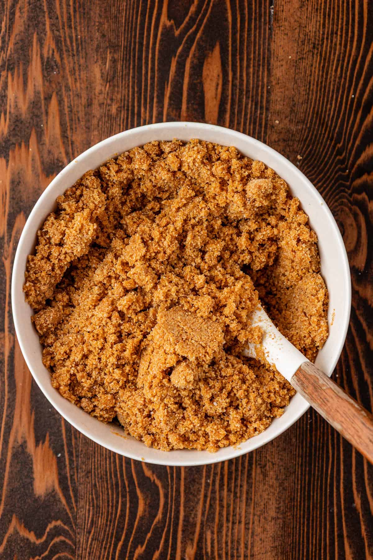 A white bowl filled with crumbly, golden brown graham cracker crust mixture sits on a dark wooden surface, with a wooden-handled spatula resting inside the bowl.