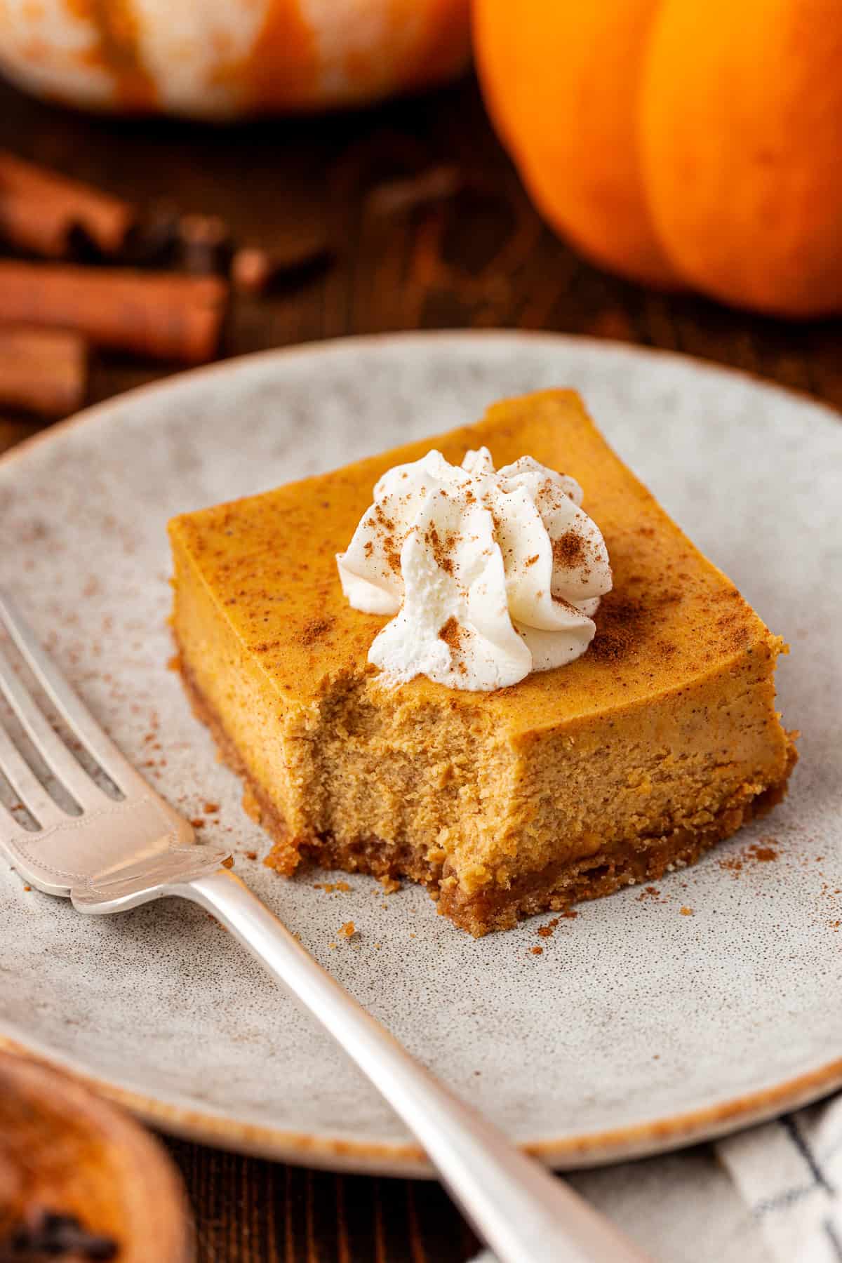 A square slice of pumpkin cheesecake with a crumbly crust, topped with a swirl of whipped cream and a sprinkle of cinnamon, sits on a plate next to a fork. Pumpkins and cinnamon sticks are in the background.