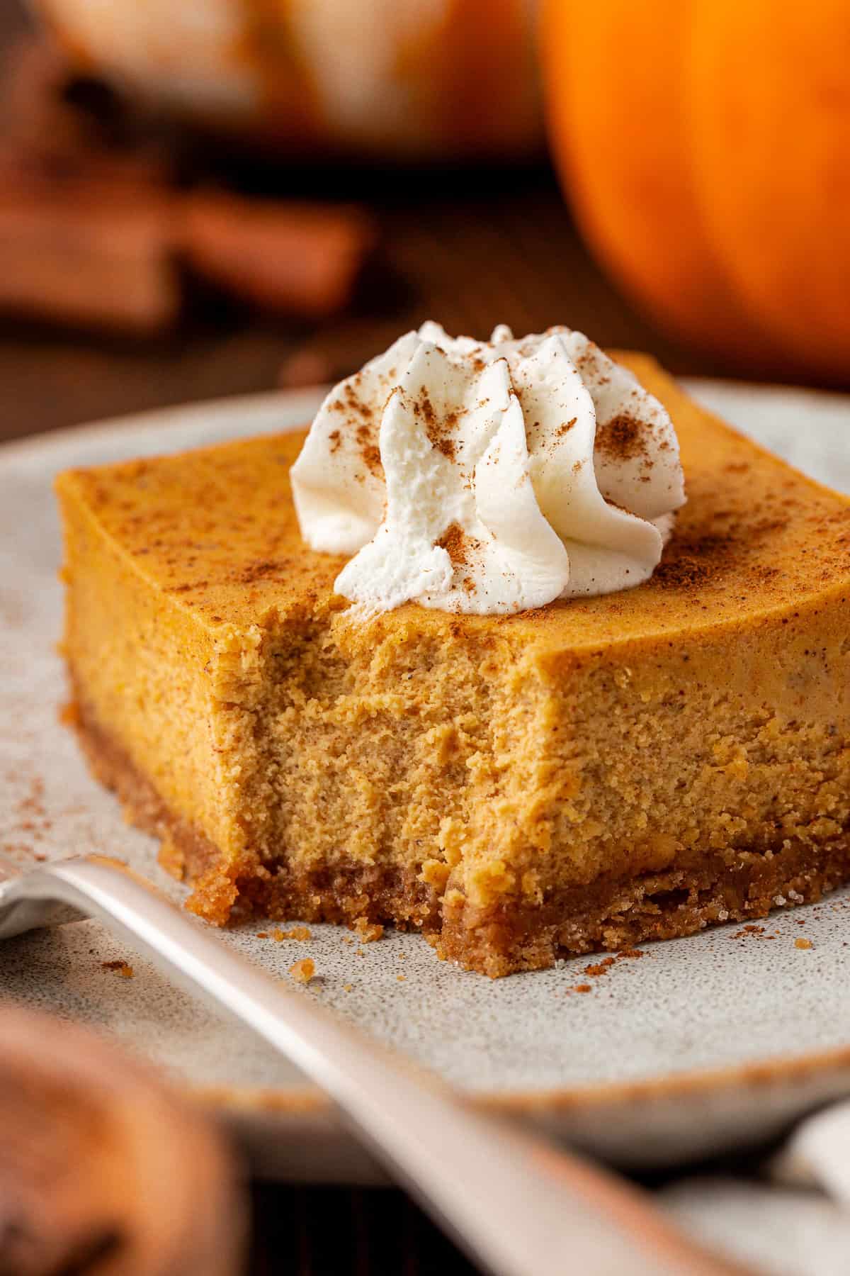 A square slice of pumpkin cheesecake with a graham cracker crust, topped with a swirl of whipped cream and sprinkled with cinnamon, sits on a plate with a fork.