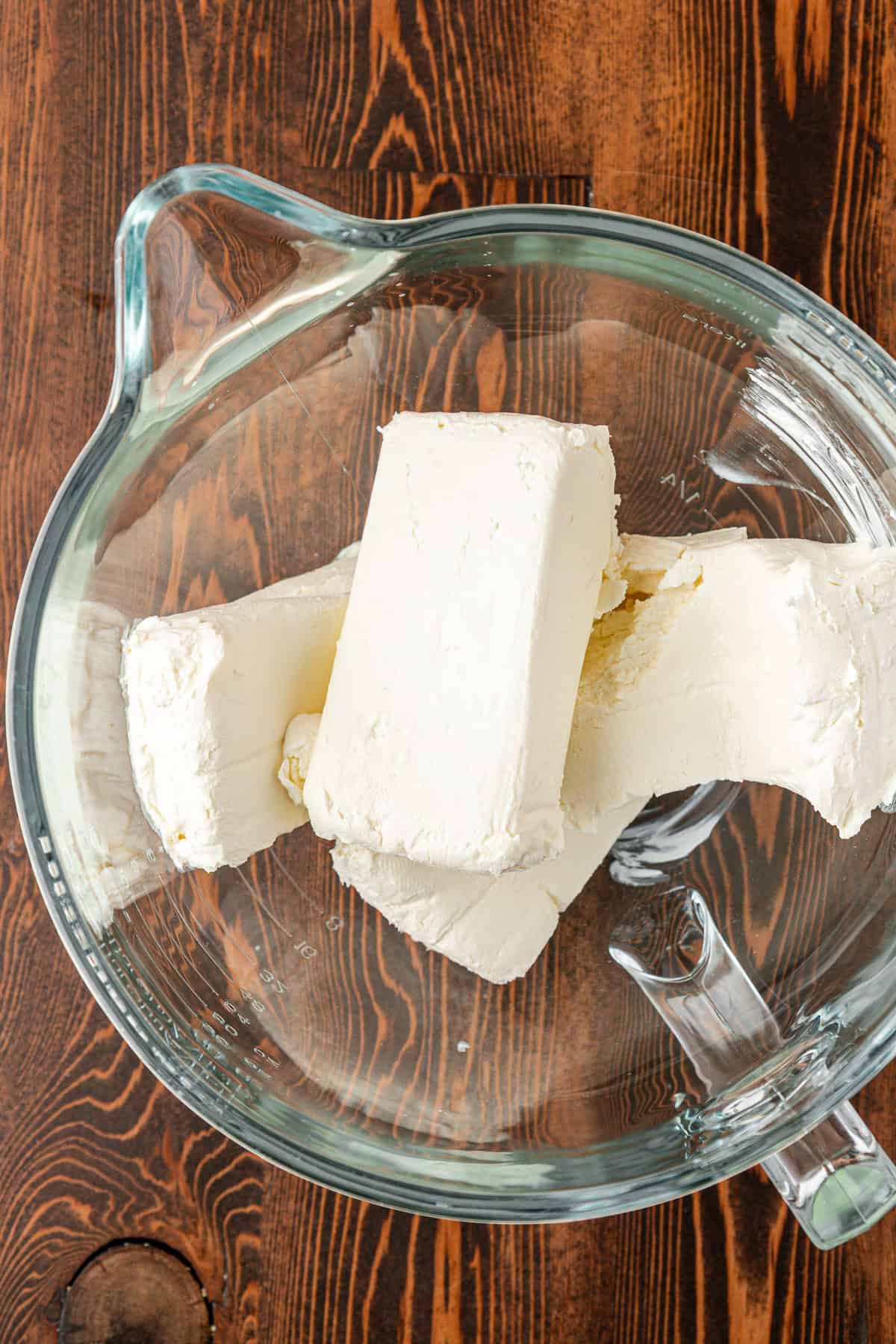 Three blocks of cream cheese in a clear glass mixing bowl, placed on a wooden surface.