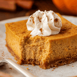 A square slice of pumpkin cheesecake with a graham cracker crust, topped with whipped cream and a sprinkle of cinnamon, sits on a plate with a bite taken out.