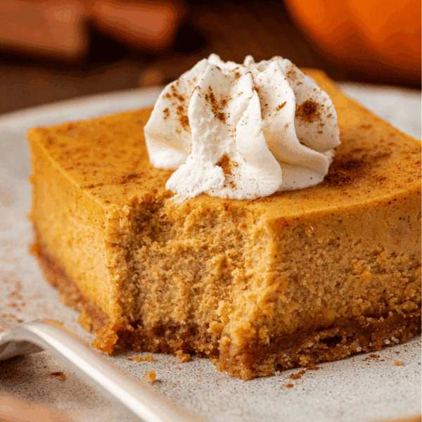 A square slice of pumpkin cheesecake with a graham cracker crust, topped with whipped cream and a sprinkle of cinnamon, sits on a plate with a bite taken out.