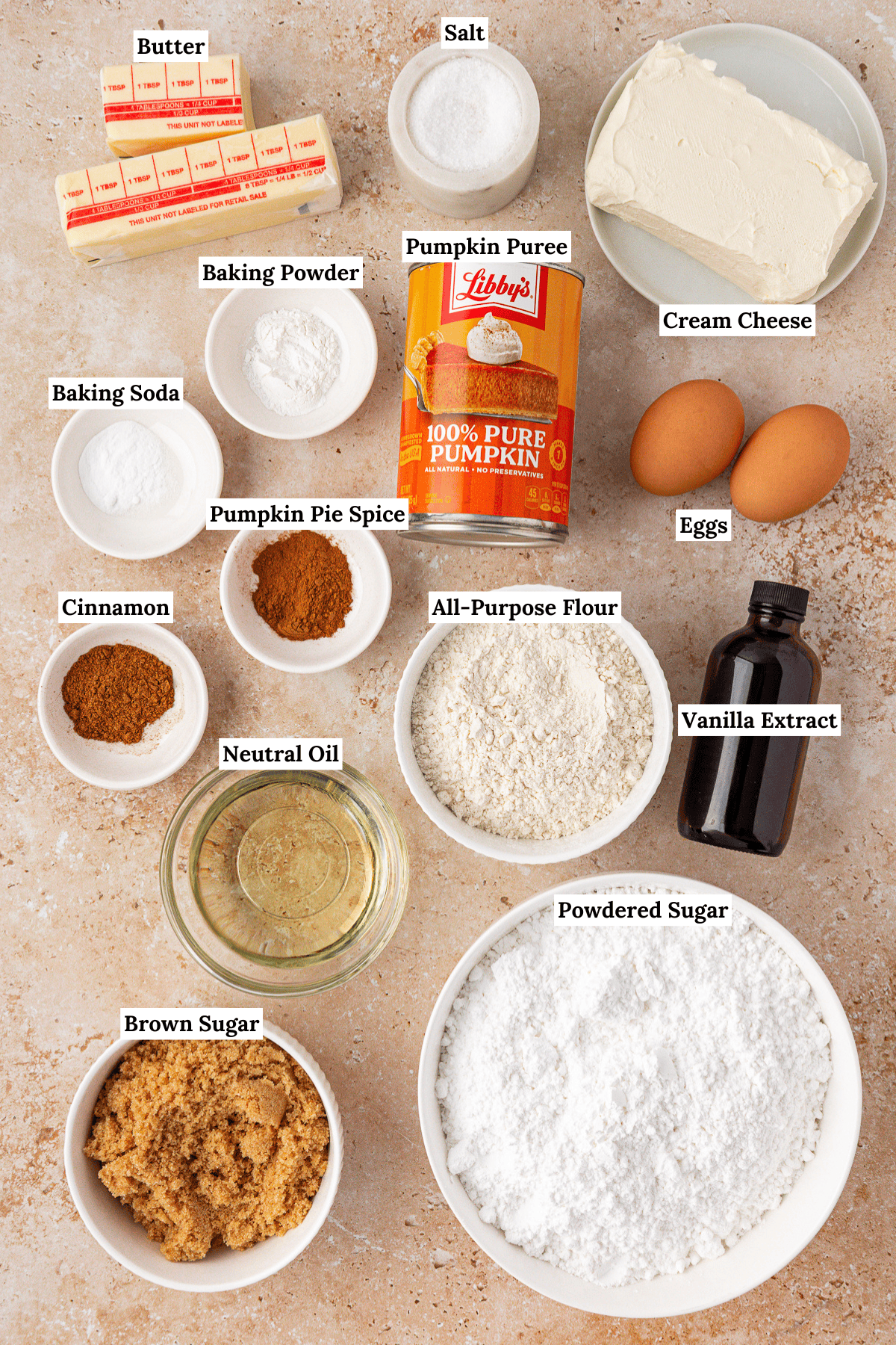 Labeled baking ingredients on a countertop, including butter, salt, cream cheese, eggs, vanilla extract, powdered sugar, brown sugar, neutral oil, all-purpose flour, pumpkin puree, pumpkin pie spice, cinnamon, baking soda, and baking powder.