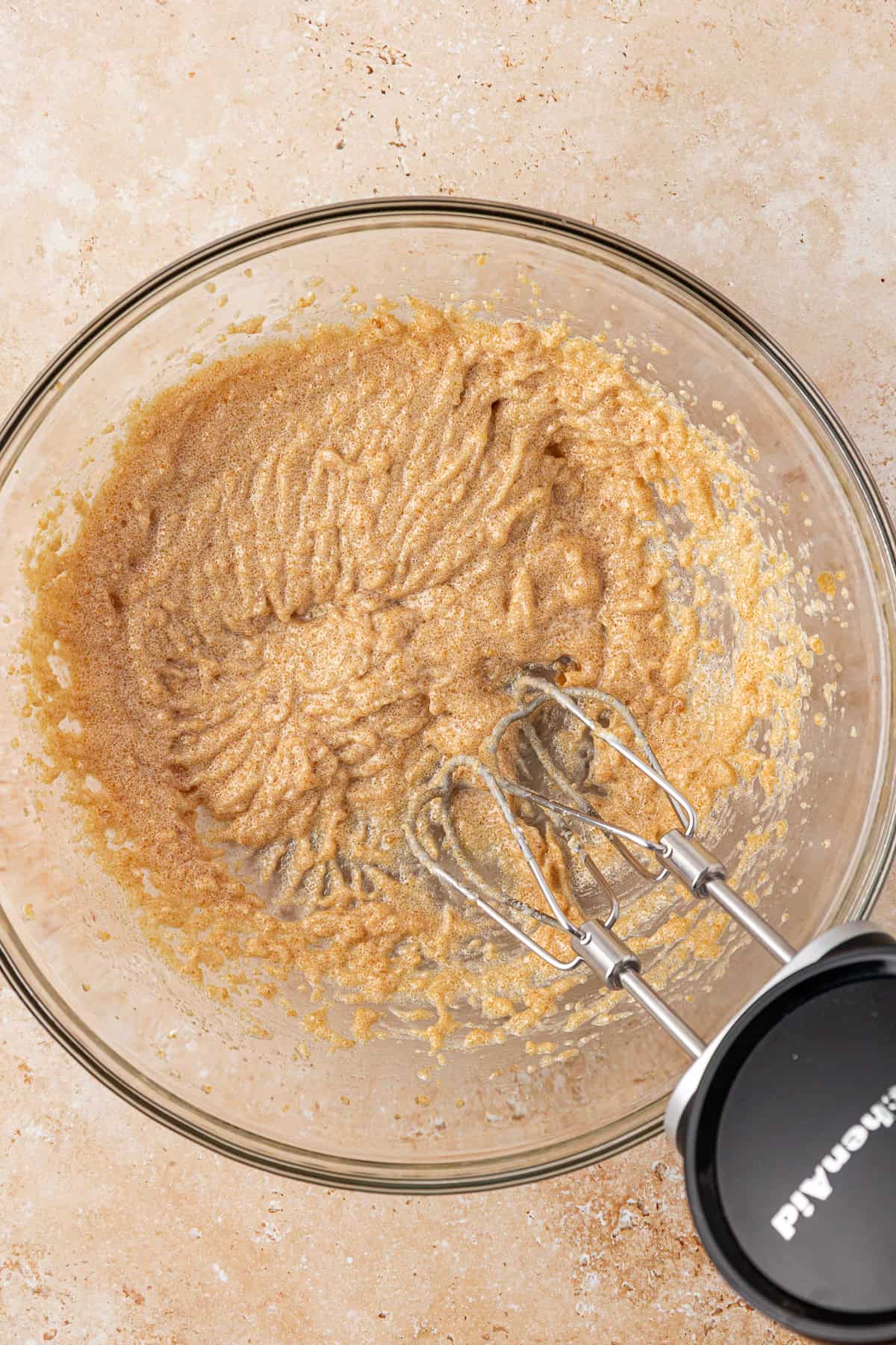 A glass mixing bowl with creamed brown sugar and butter, partially mixed with an electric hand mixer resting inside the bowl on a beige countertop.