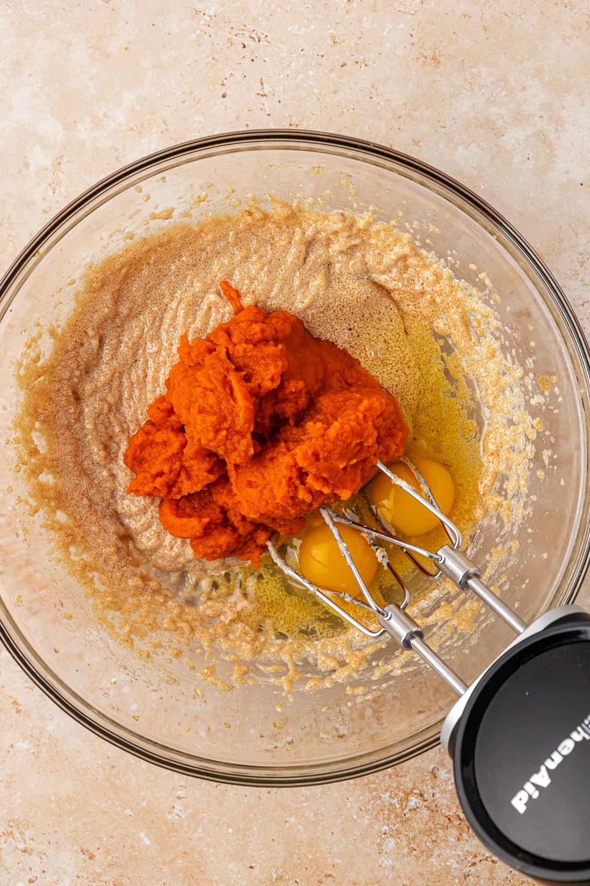 A glass bowl with mixed brown sugar and butter, three raw eggs, and a mound of pumpkin puree, with a hand mixer positioned inside, on a beige countertop.