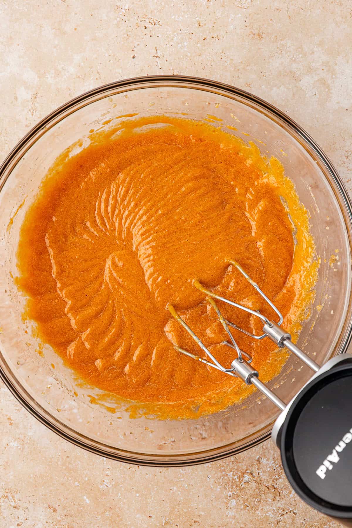 A glass mixing bowl filled with orange batter sits on a light countertop. An electric hand mixer with two beaters rests inside the bowl, showing swirled patterns in the smooth batter.