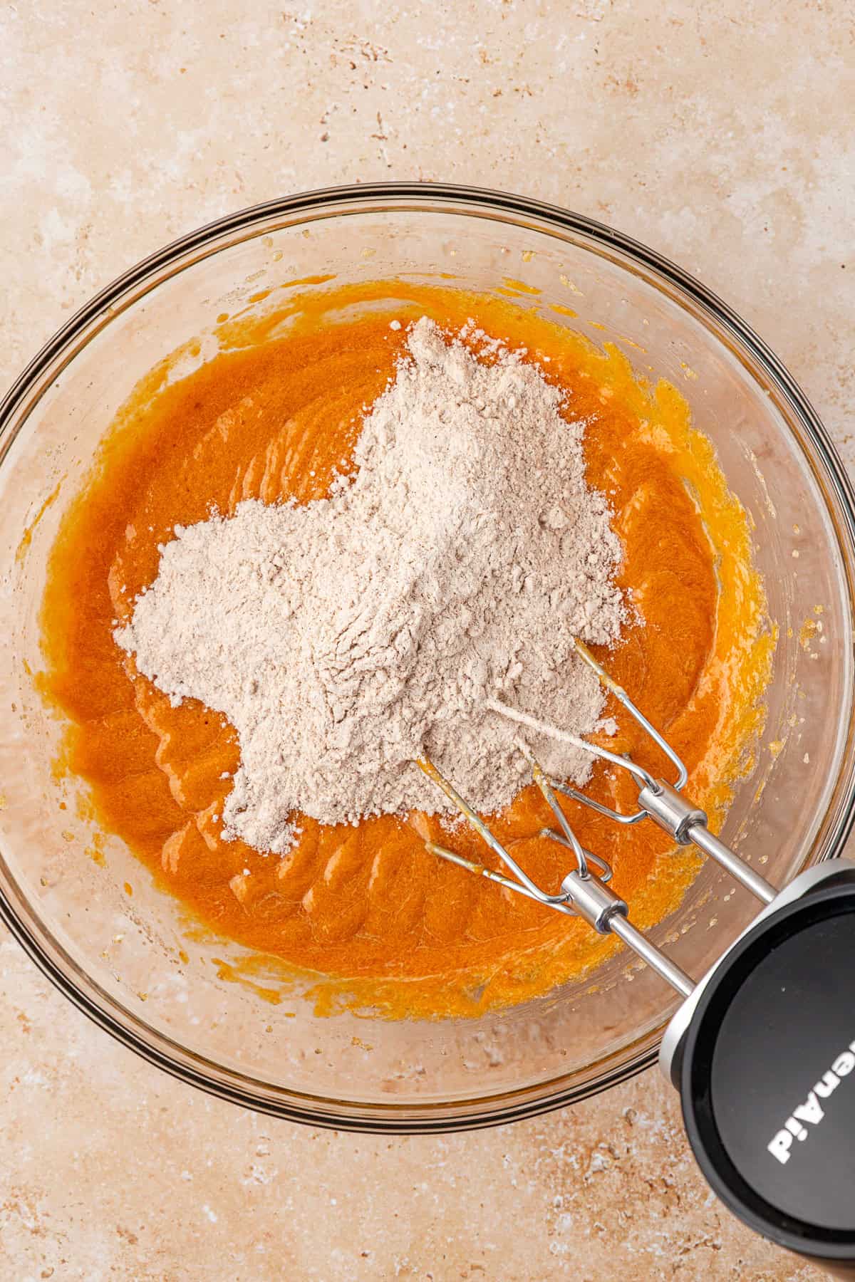 A glass bowl with orange batter and a mound of flour on top, partially mixed with an electric hand mixer resting inside the bowl, on a light-colored countertop.