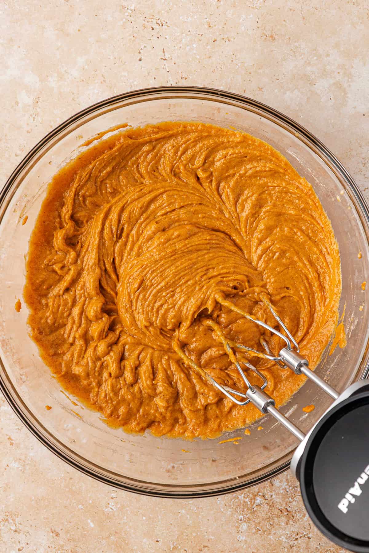A glass bowl filled with pumpkin-colored batter being mixed with an electric hand mixer on a light brown countertop.