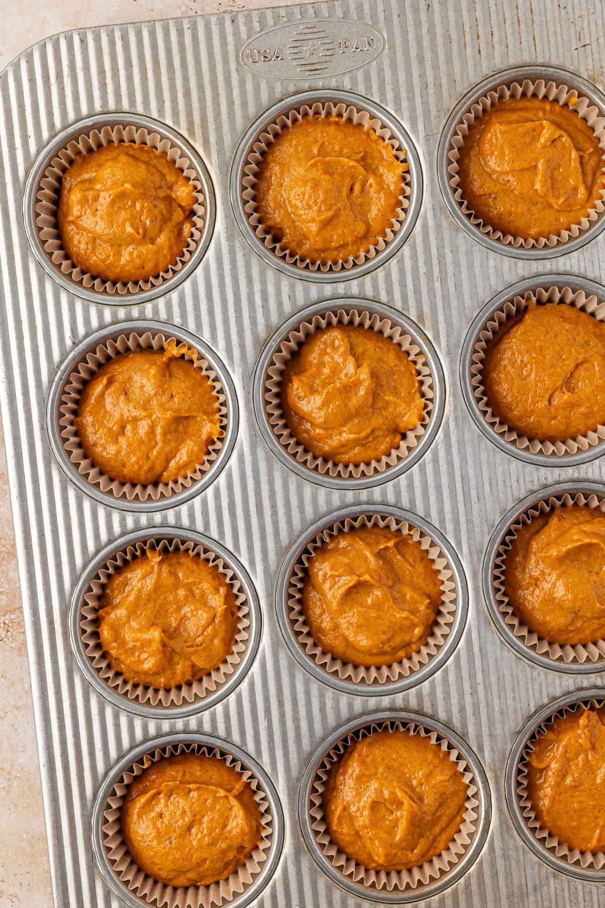 A metal muffin tray filled with twelve paper liners, each containing unbaked orange muffin batter. The tray is on a light-colored surface.