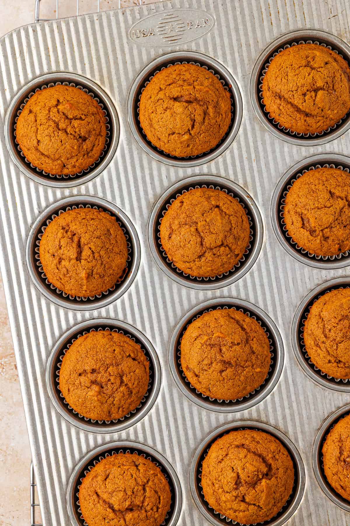 A muffin tin filled with freshly baked golden-brown muffins, each in a black paper liner, resting on a cooling rack.