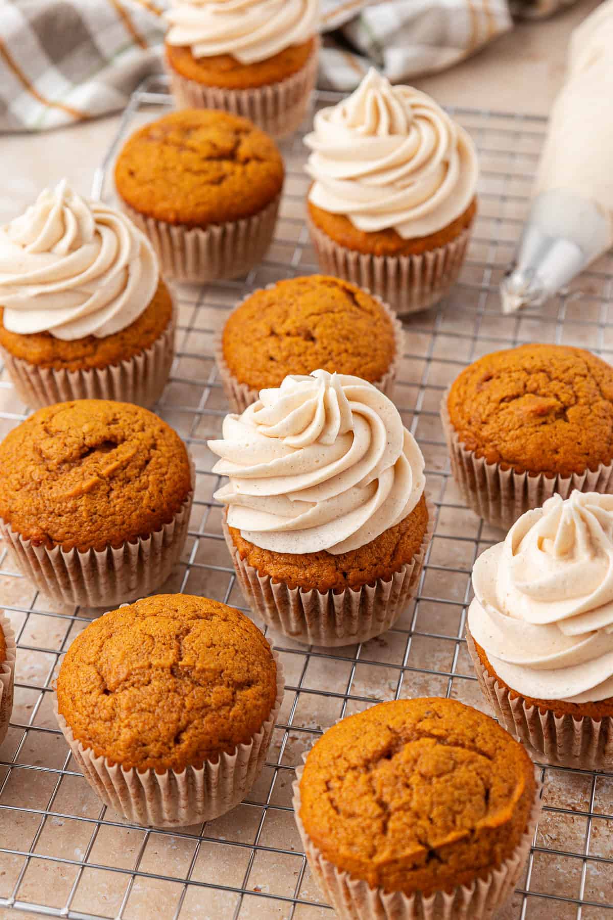 Pumpkin cupcakes on a cooling rack, some topped with swirls of cream-colored frosting and others plain. A piping bag with frosting is visible in the background.