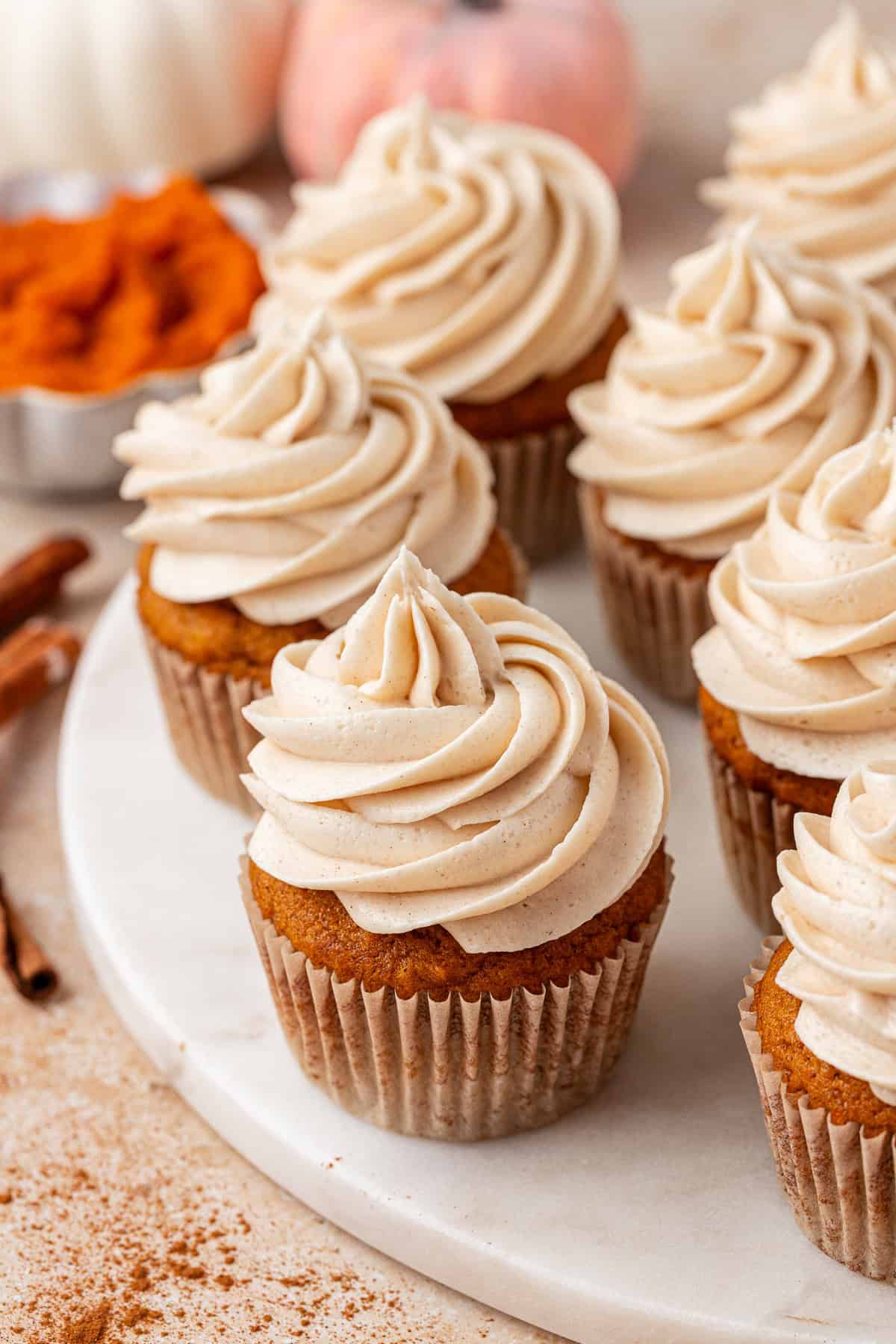 Pumpkin cupcakes with swirled cinnamon frosting sit on a white marble surface, surrounded by cinnamon sticks and a bowl of pumpkin puree in the background.