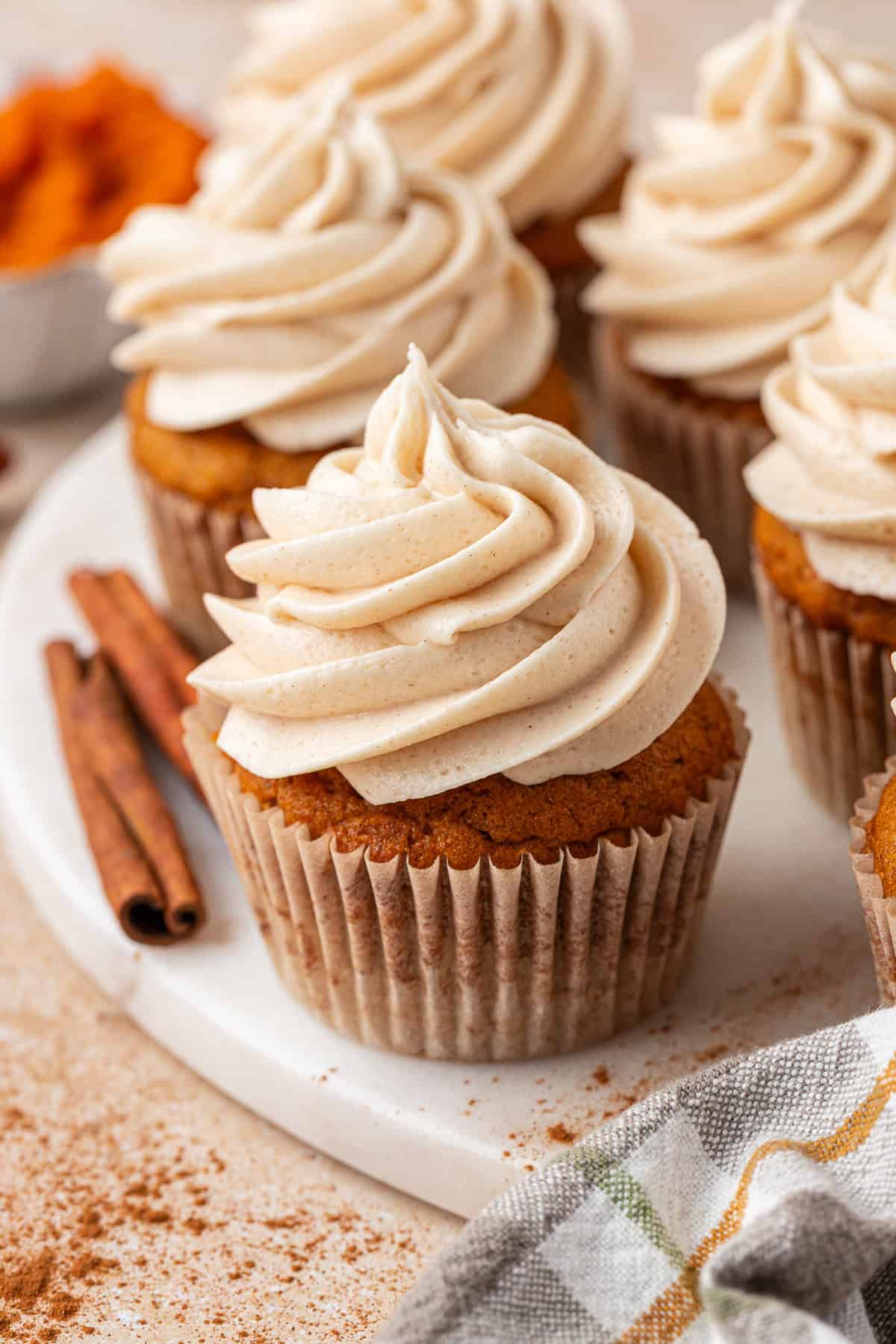 A close-up of pumpkin cupcakes with swirls of creamy frosting on top, arranged on a white plate. Two cinnamon sticks and a sprinkle of cinnamon powder are nearby. A checkered napkin and a bowl of orange filling are in the background.