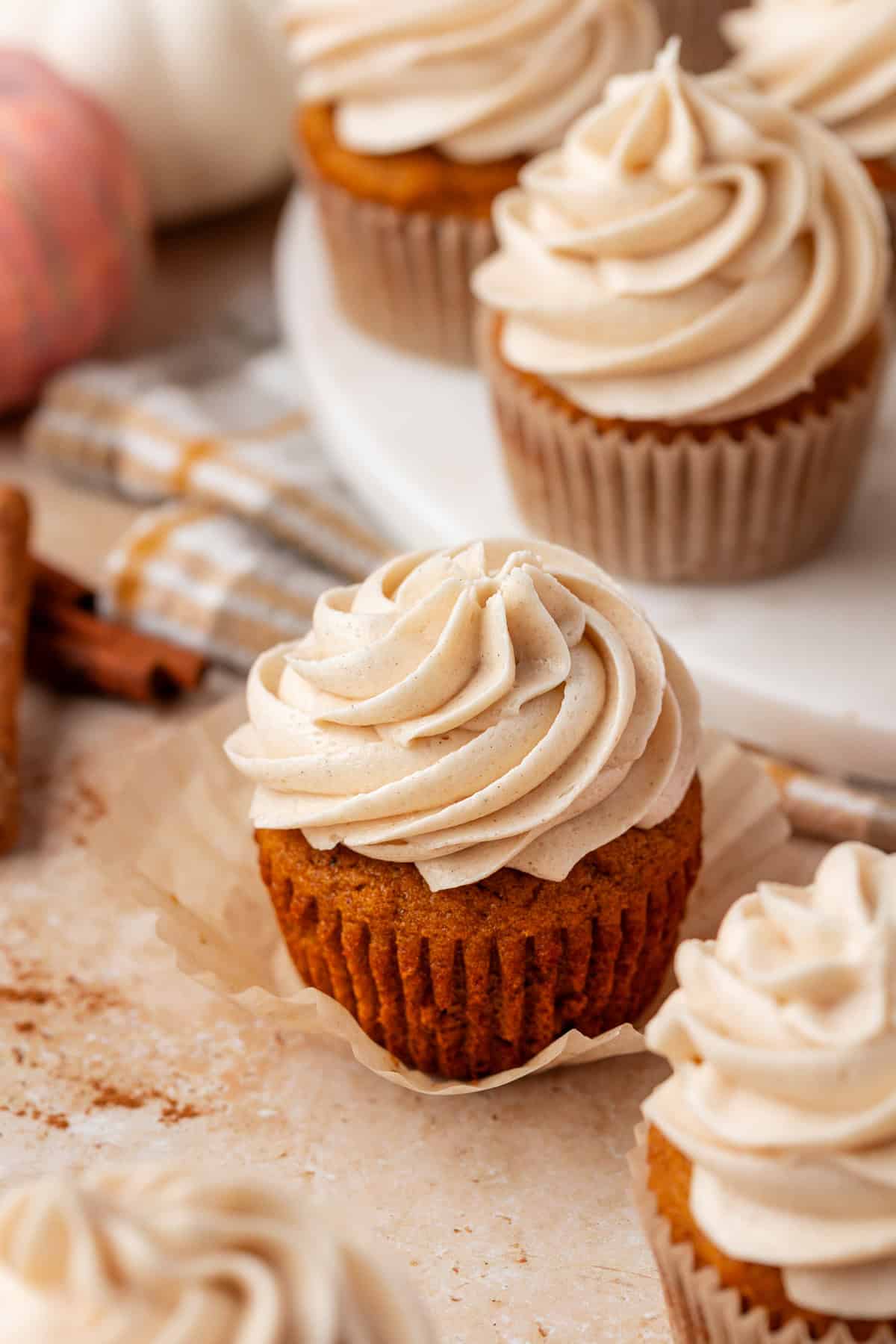 A close-up of pumpkin cupcakes topped with swirls of creamy frosting, set on a light surface with fall-themed decor and cinnamon sticks in the background.