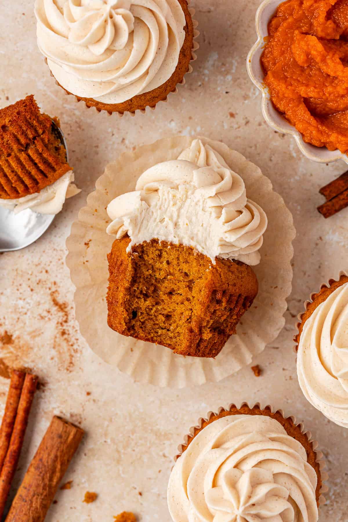 A pumpkin cupcake with swirled frosting sits on a wrapper, showing a bite taken out. Surrounding it are whole cupcakes, a bowl of pumpkin puree, cinnamon sticks, and scattered crumbs on a light surface.