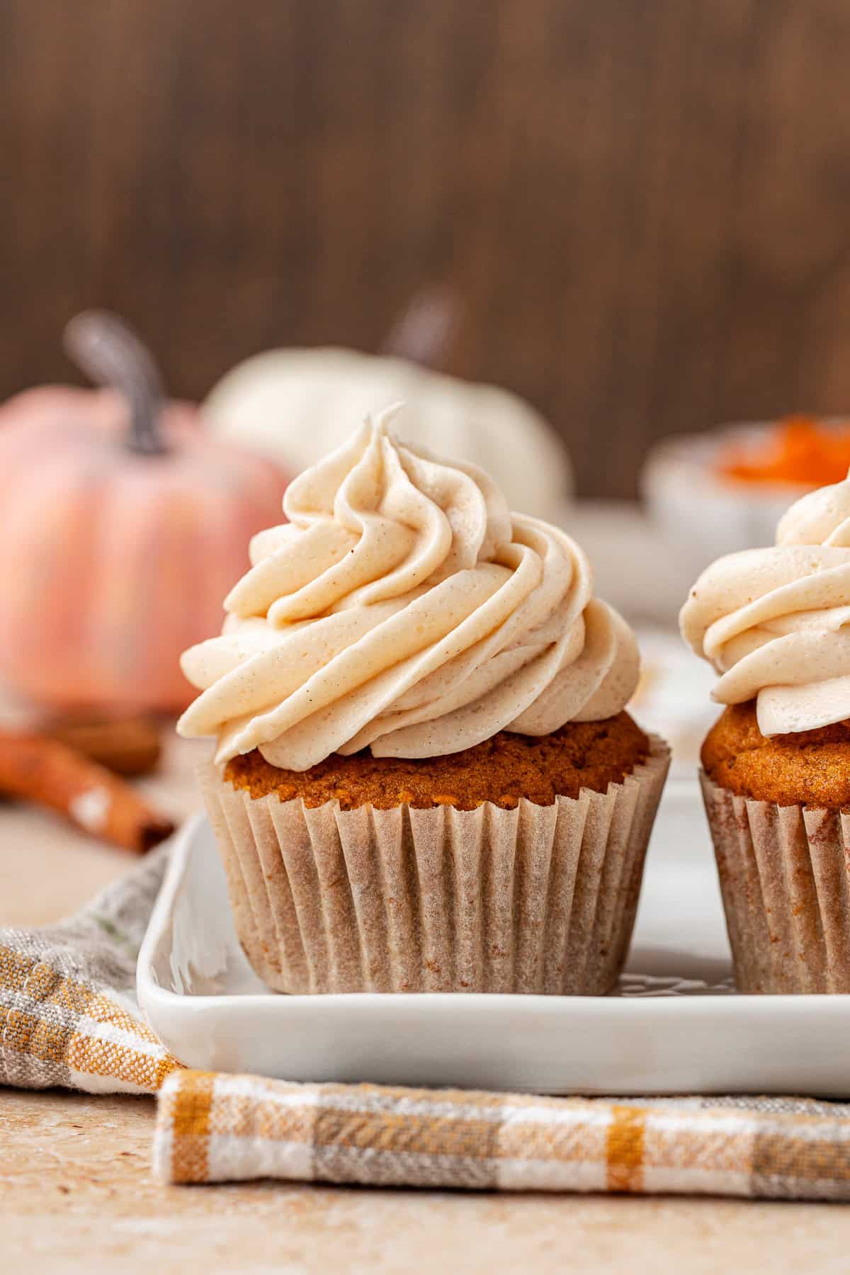 A pumpkin cupcake topped with swirled cream frosting sits on a white plate, with another cupcake beside it. A plaid napkin, cinnamon sticks, and blurred pumpkins are in the background.