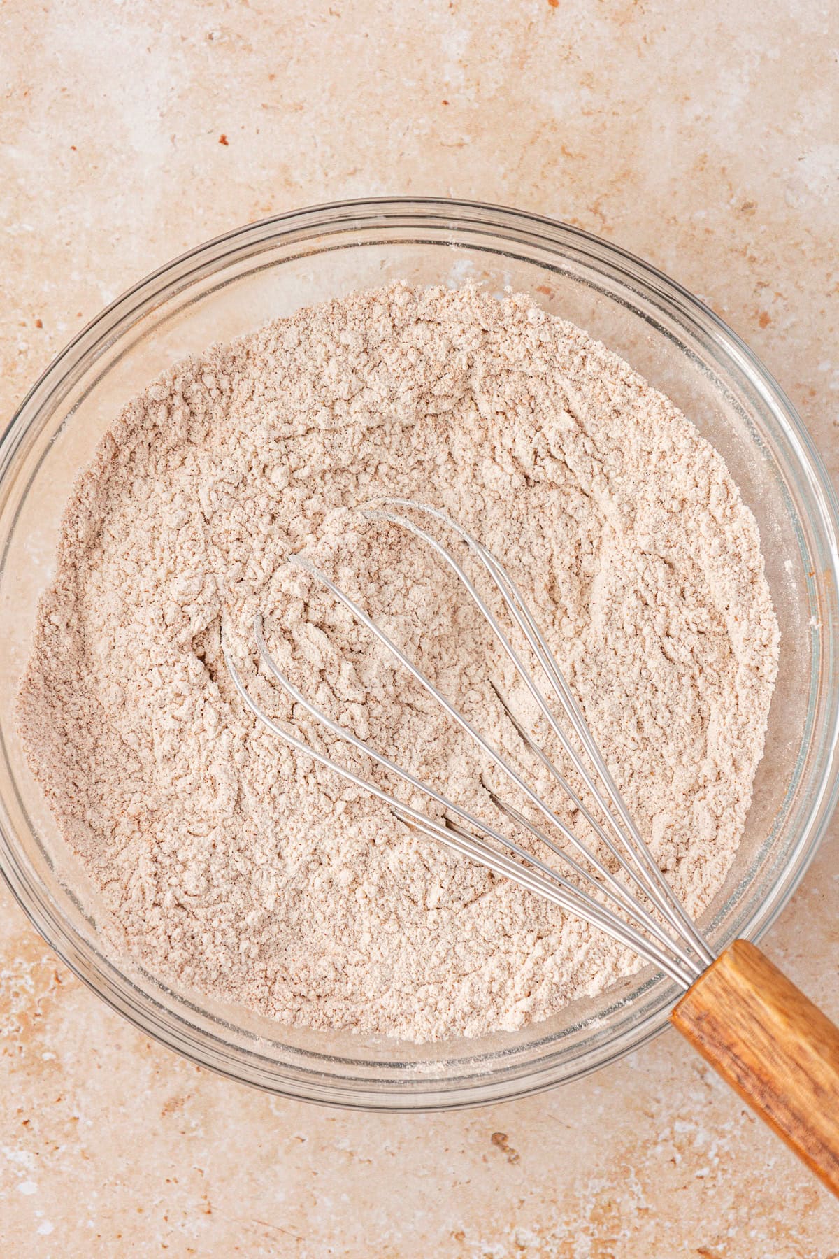 A glass bowl filled with a light brown flour mixture and a metal whisk with a wooden handle, resting on a light beige countertop.