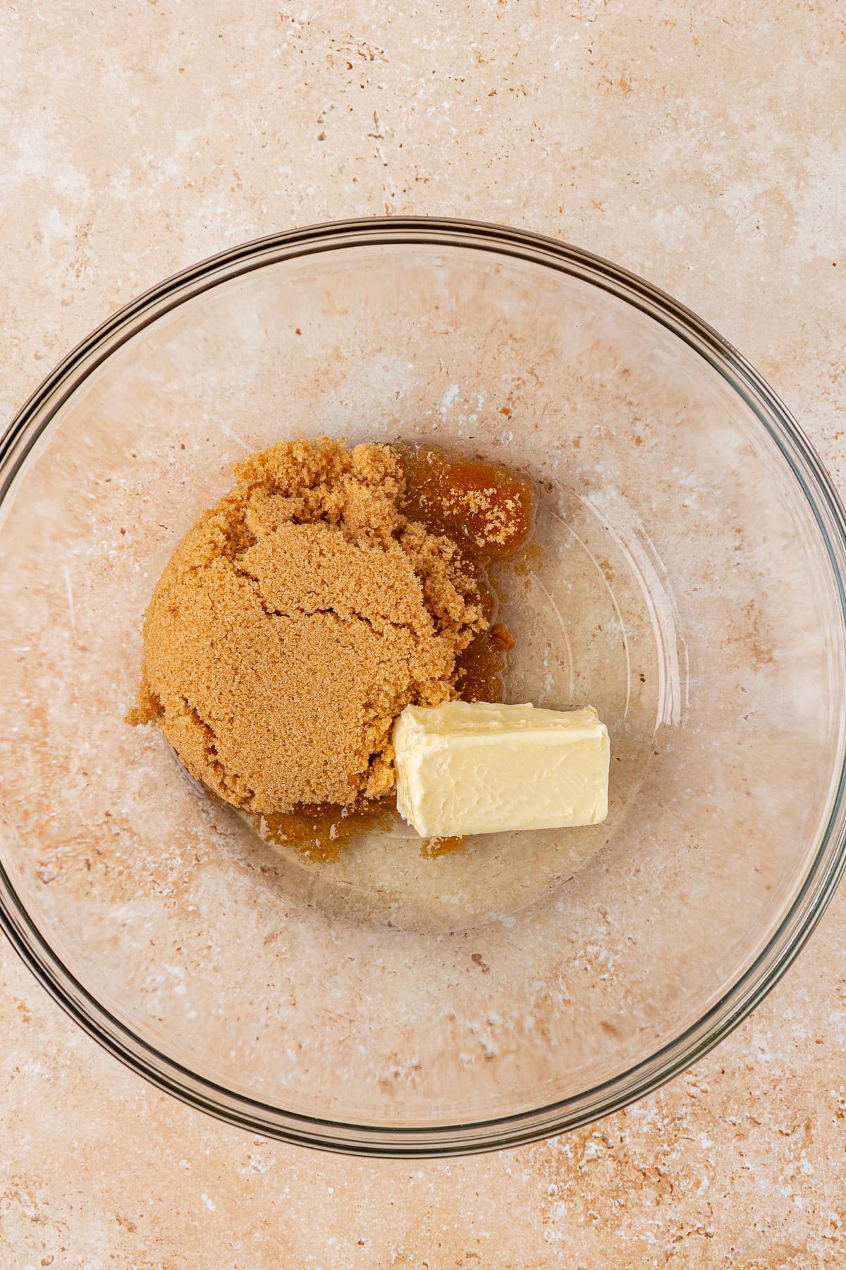 A glass mixing bowl containing a mound of brown sugar and a chunk of butter on a light beige countertop.