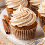 A pumpkin cupcake with swirled cinnamon frosting sits on a white plate, with cinnamon sticks nearby and other cupcakes in the background.