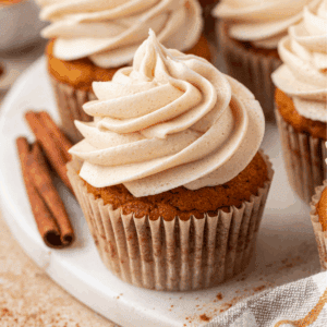 A pumpkin cupcake with swirled cinnamon frosting sits on a white plate, with cinnamon sticks nearby and other cupcakes in the background.
