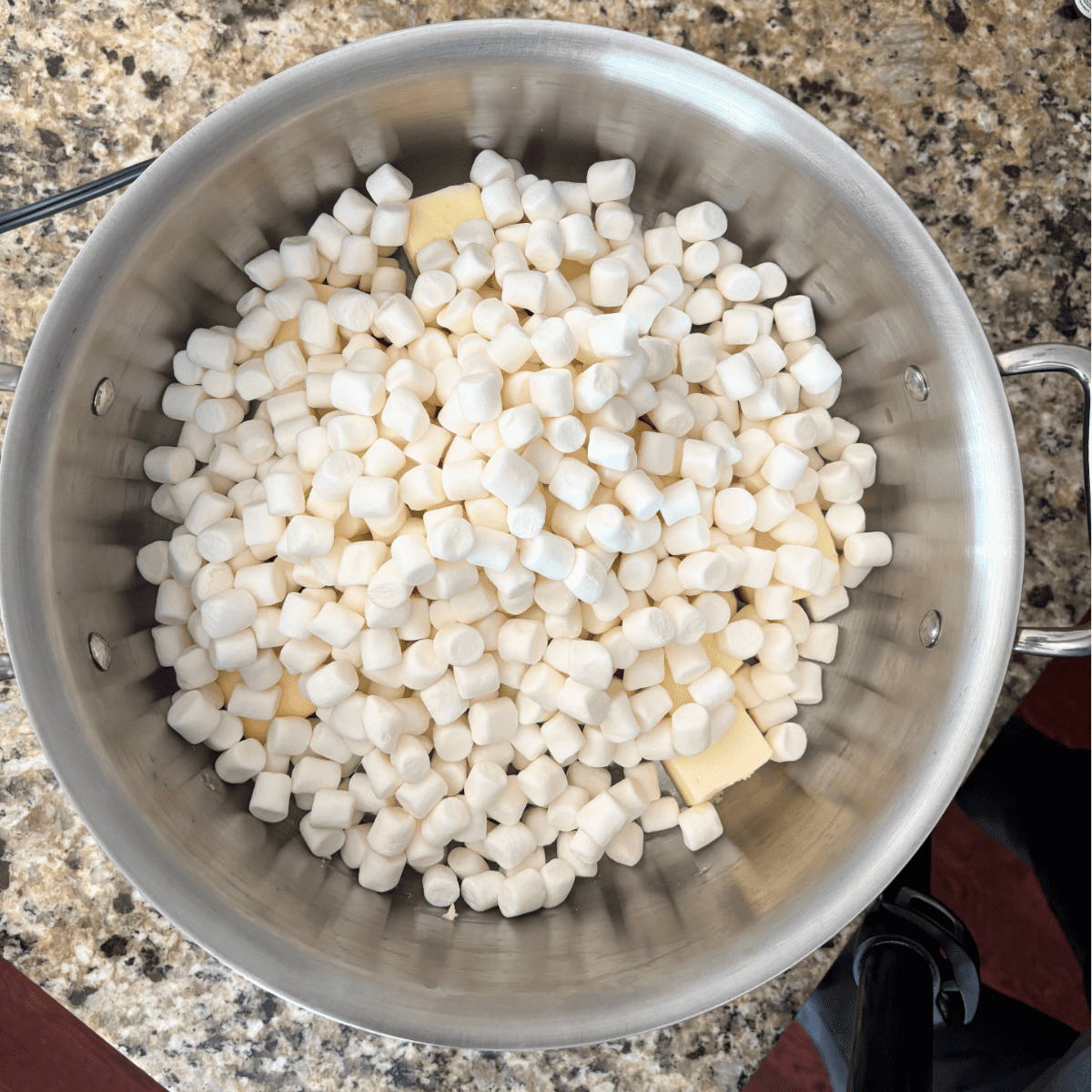 A large silver pot on a granite countertop filled with mini marshmallows and pieces of butter, ready to be melted together.