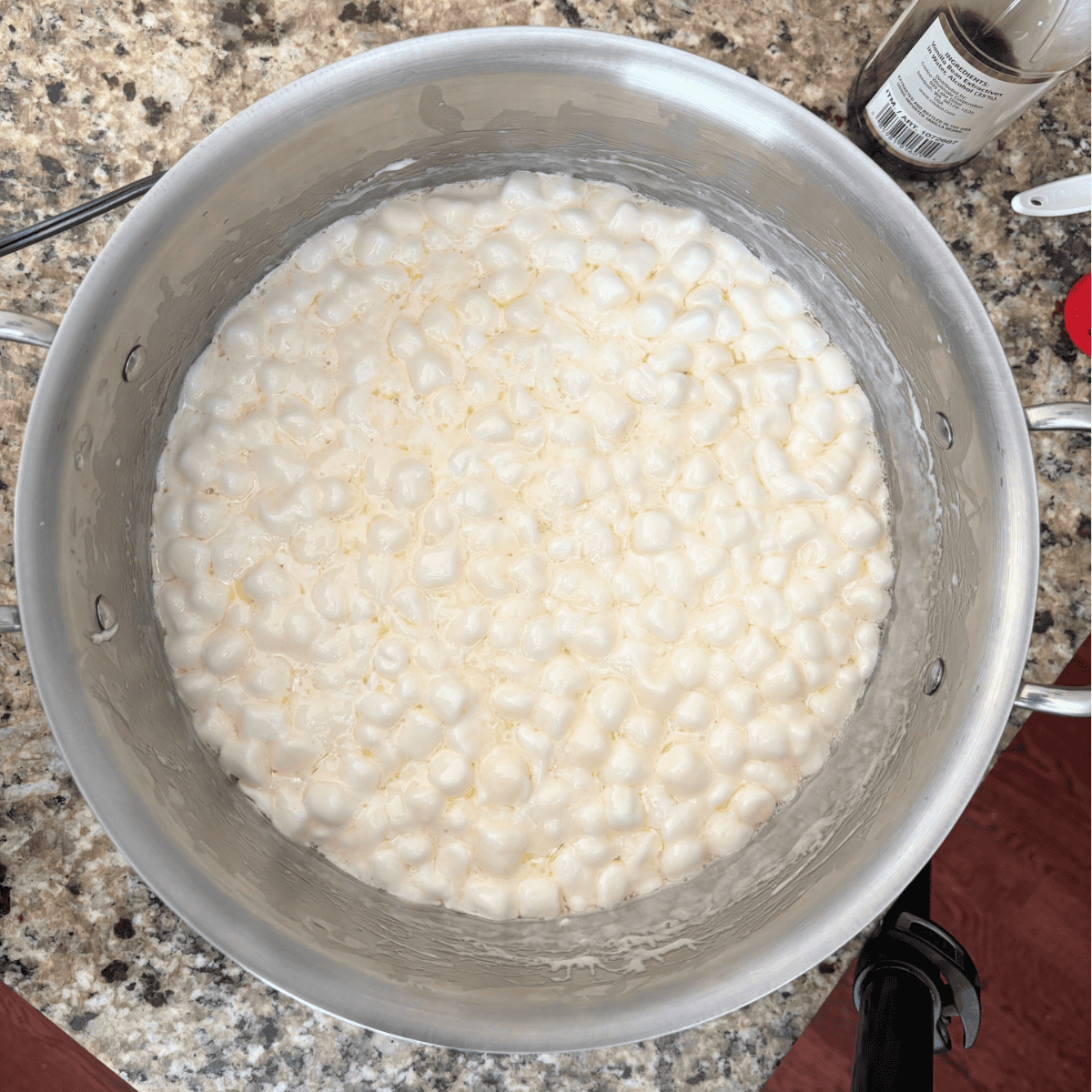 A large silver pot on a granite countertop containing melted mini marshmallows. A bottle of vanilla extract is visible nearby.