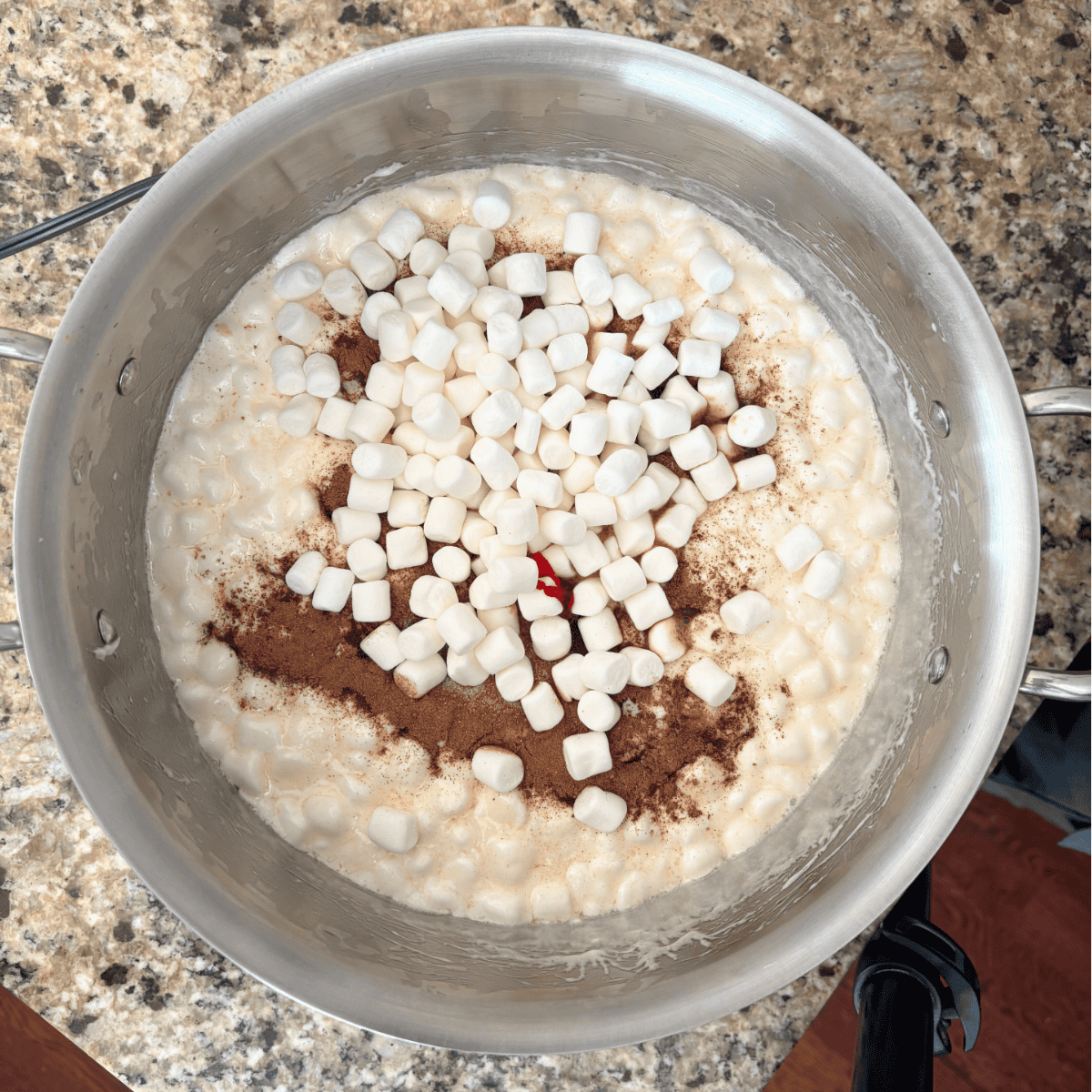 A pot on a granite countertop filled with melted marshmallows, mini marshmallows, cinnamon, and a drop of red food coloring, ready to be mixed.