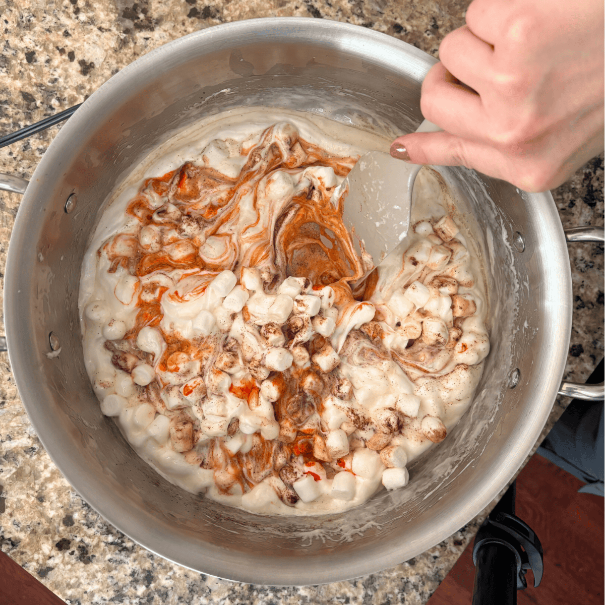 A hand holding a spatula stirs mini marshmallows with melted butter and swirls of orange coloring in a large metal pot on a granite countertop.