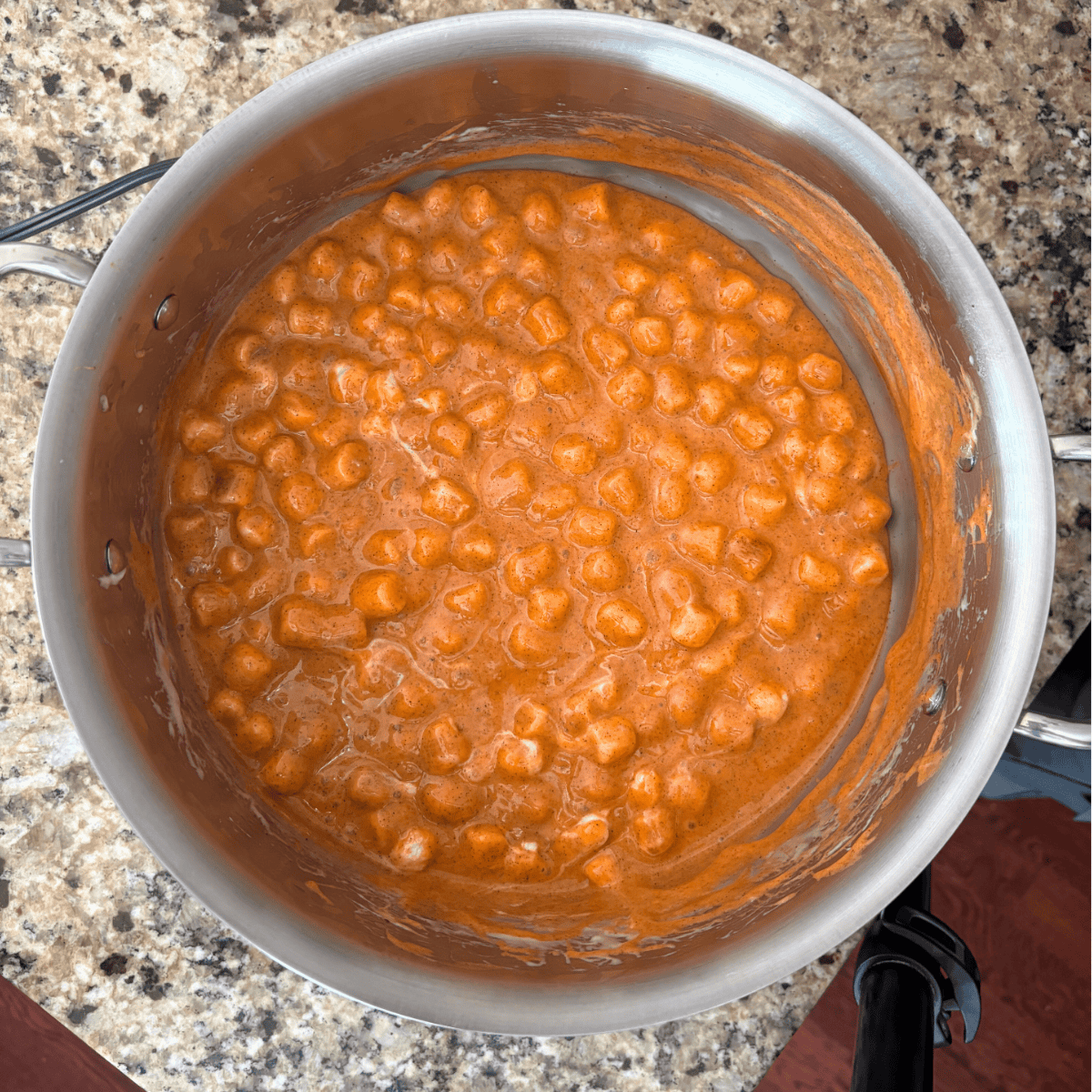 A stainless steel pot filled with mini marshmallows melting into a thick, orange sauce sits on a speckled granite countertop. The mixture appears creamy and smooth, partially coating the sides of the pot.