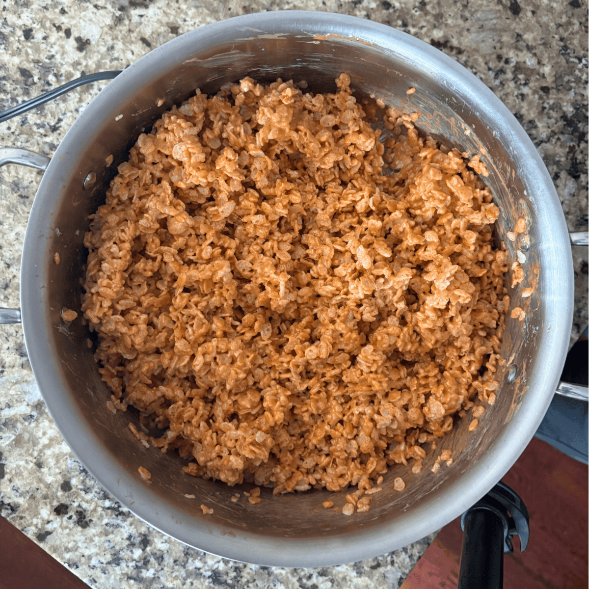 A stainless steel pot on a granite countertop filled with crispy rice cereal mixed with melted chocolate and possibly peanut butter or another brown ingredient.