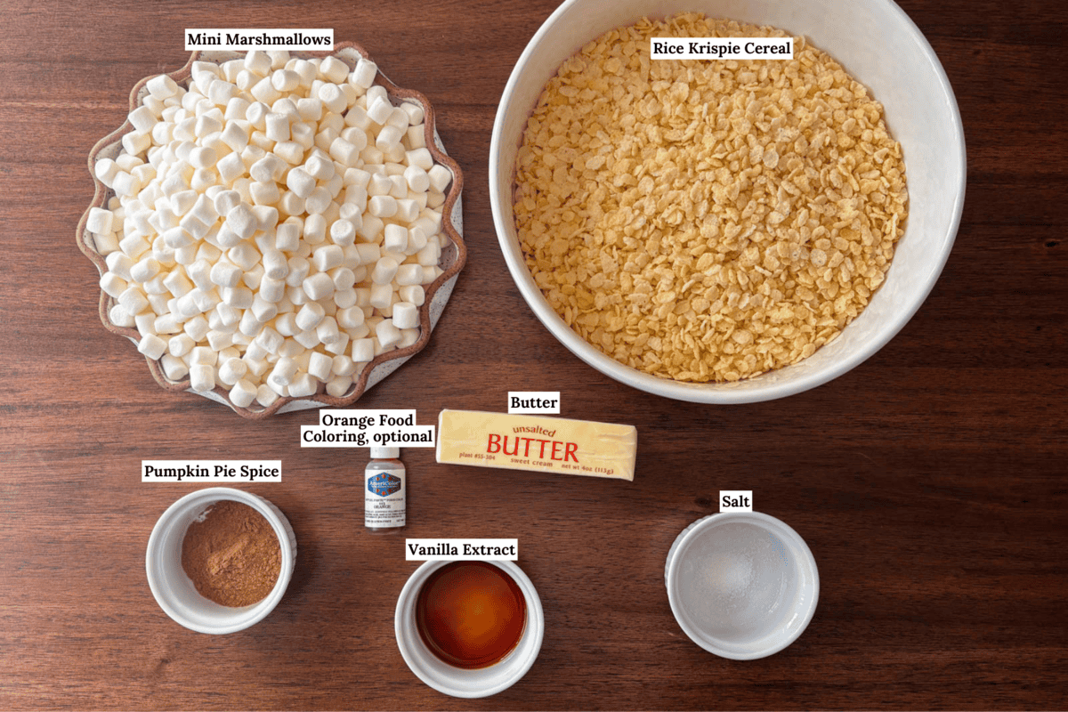 Ingredients for a recipe on a wooden table: a bowl of mini marshmallows, a bowl of Rice Krispie cereal, pumpkin pie spice, vanilla extract, butter, salt, and orange food coloring. Each item is labeled.