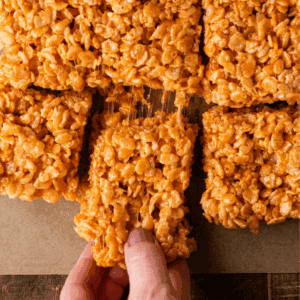 A hand lifts a gooey rice crispy treat from a batch of square-cut treats on parchment paper. The treats are golden brown with visible puffed rice cereal, stuck together with melted marshmallow.