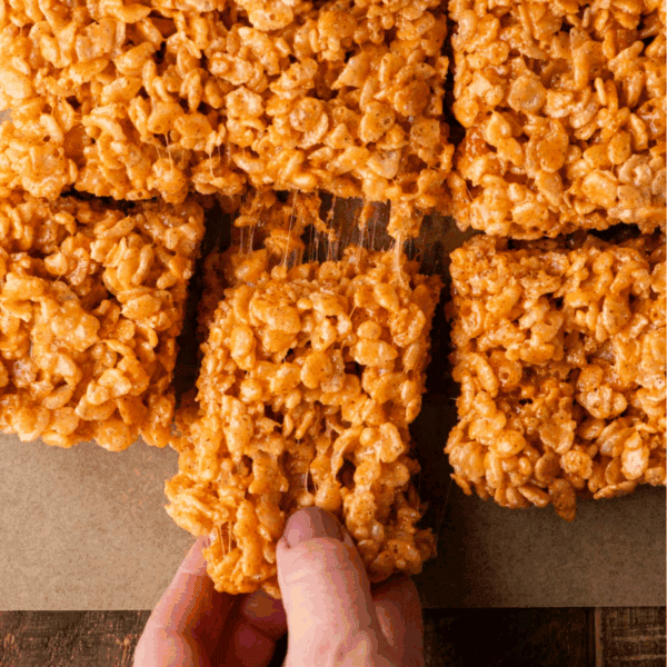 A hand lifts a gooey rice crispy treat from a batch of square-cut treats on parchment paper. The treats are golden brown with visible puffed rice cereal, stuck together with melted marshmallow.