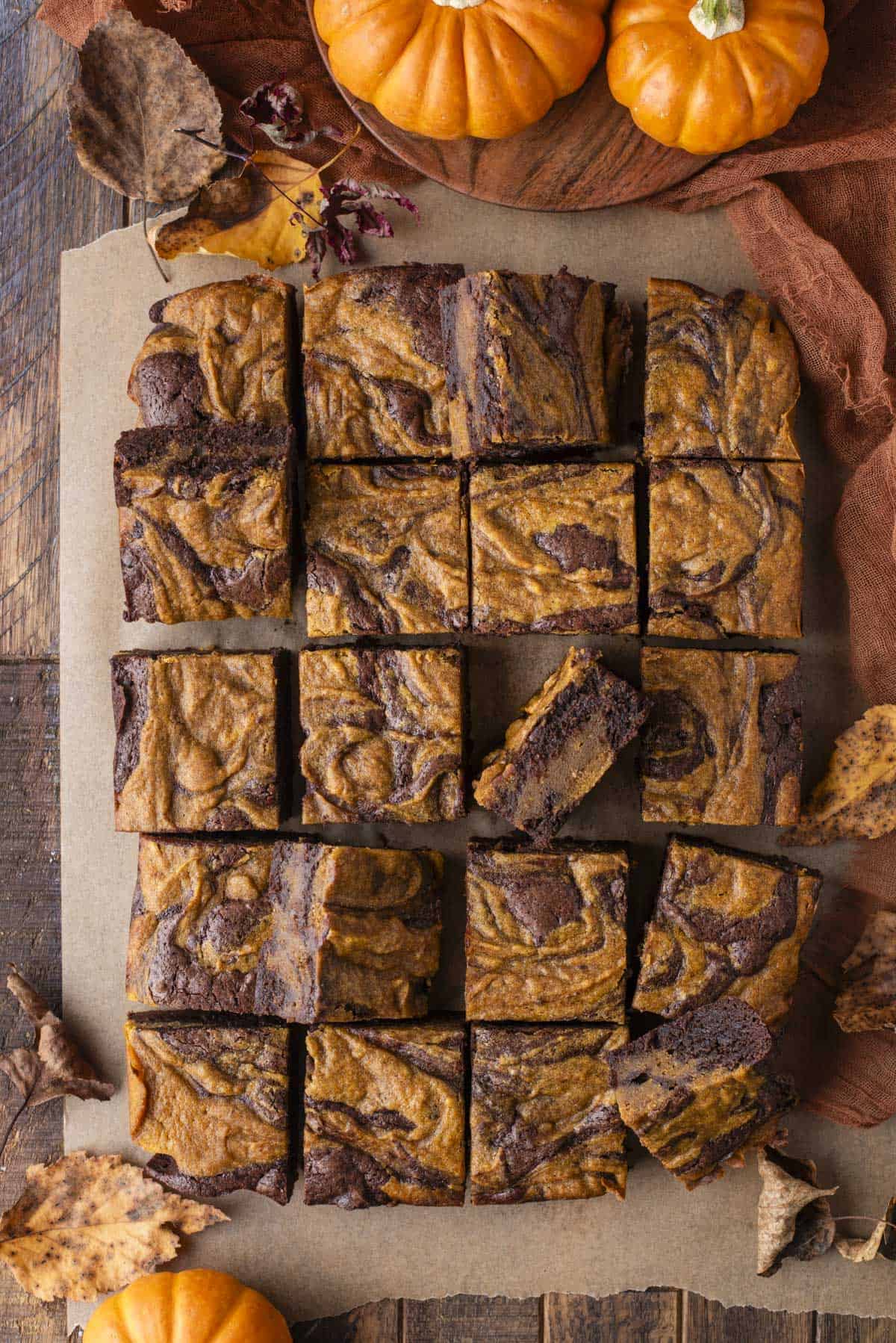 Top-down view of pumpkin swirl brownies cut into squares on parchment paper, surrounded by autumn leaves, mini pumpkins, and a brown cloth, set on a rustic wooden table.