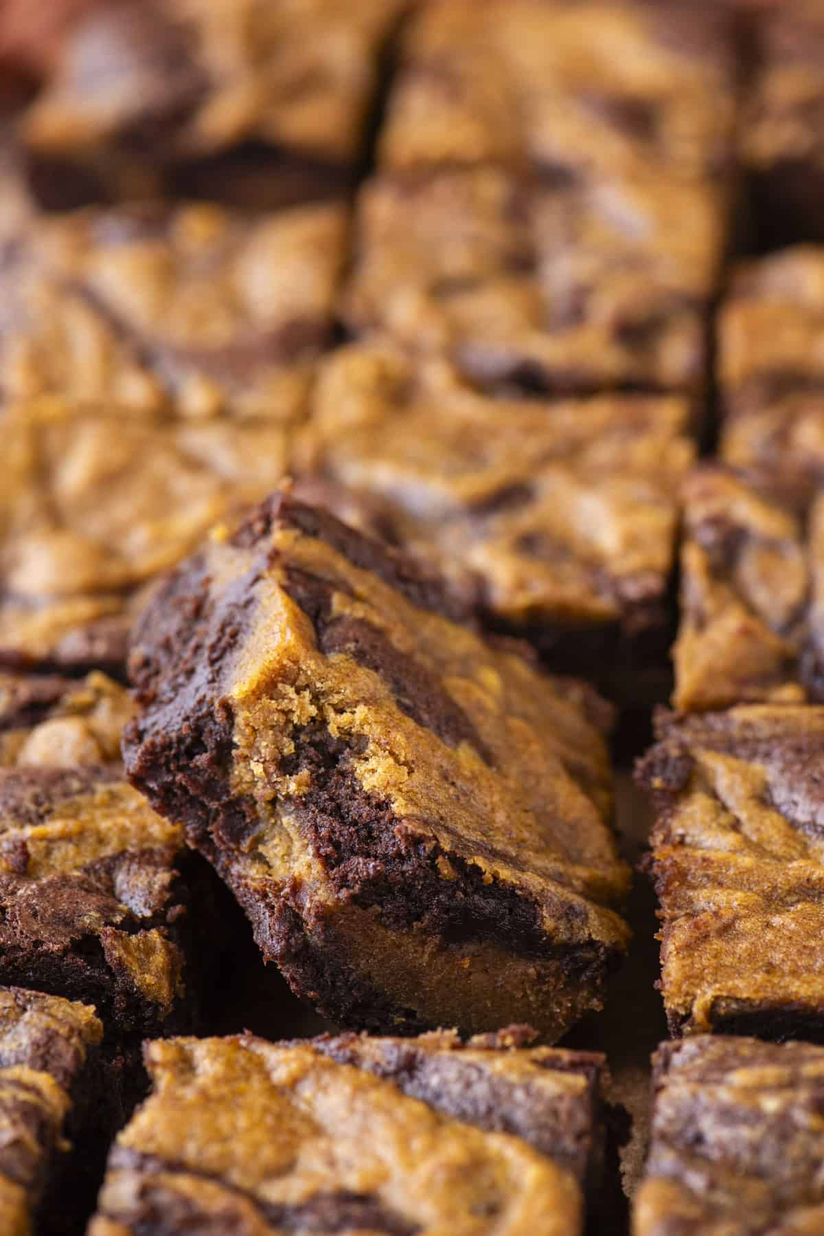 Close-up of several square brownies with a marbled chocolate and peanut butter swirl pattern, showing their rich, fudgy texture and golden brown tops. One brownie is slightly raised above the others.