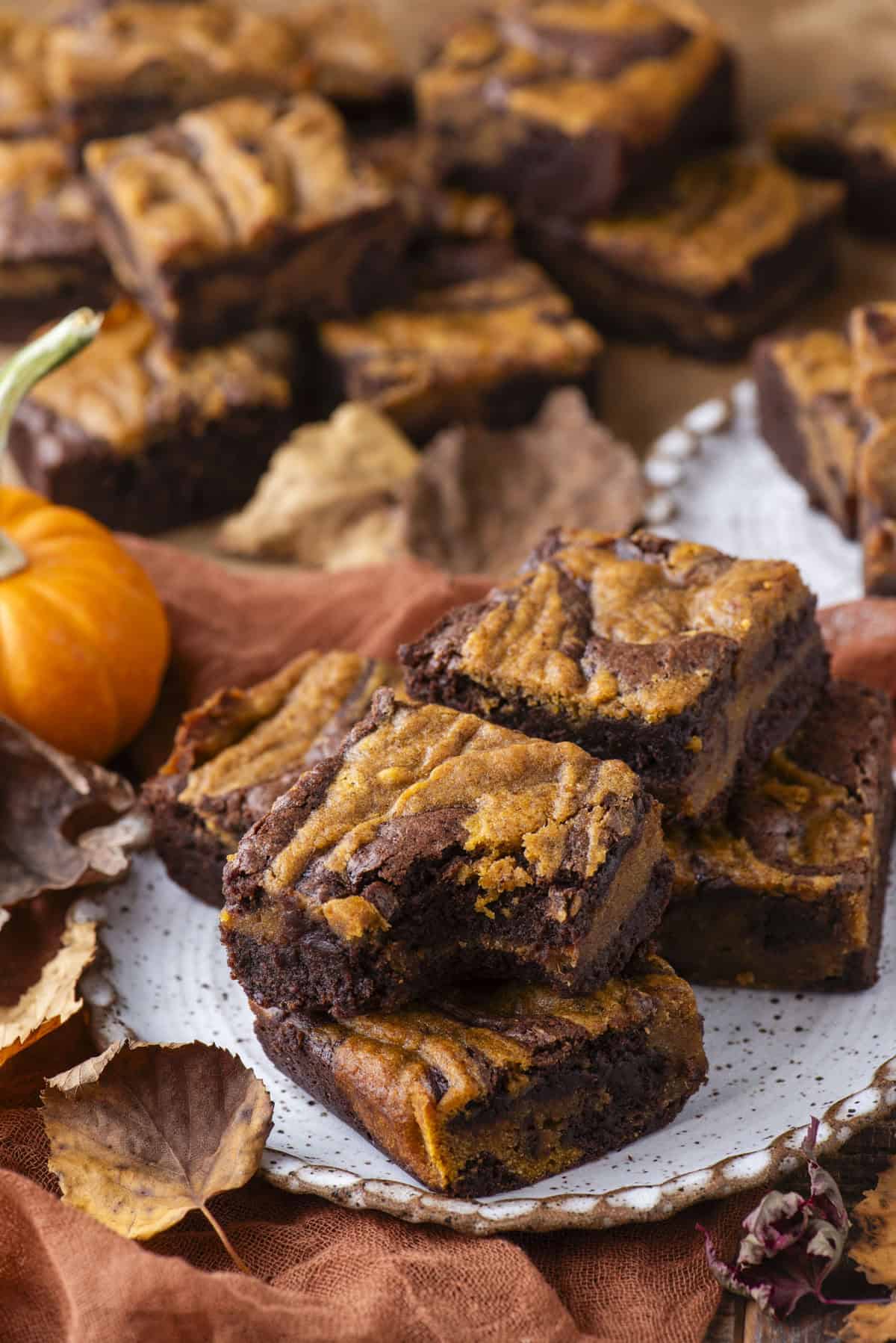 A plate of pumpkin swirl brownies sits on a brown cloth, surrounded by scattered autumn leaves and a small pumpkin, with more brownies in the background. The brownies feature a marbled orange and chocolate pattern.