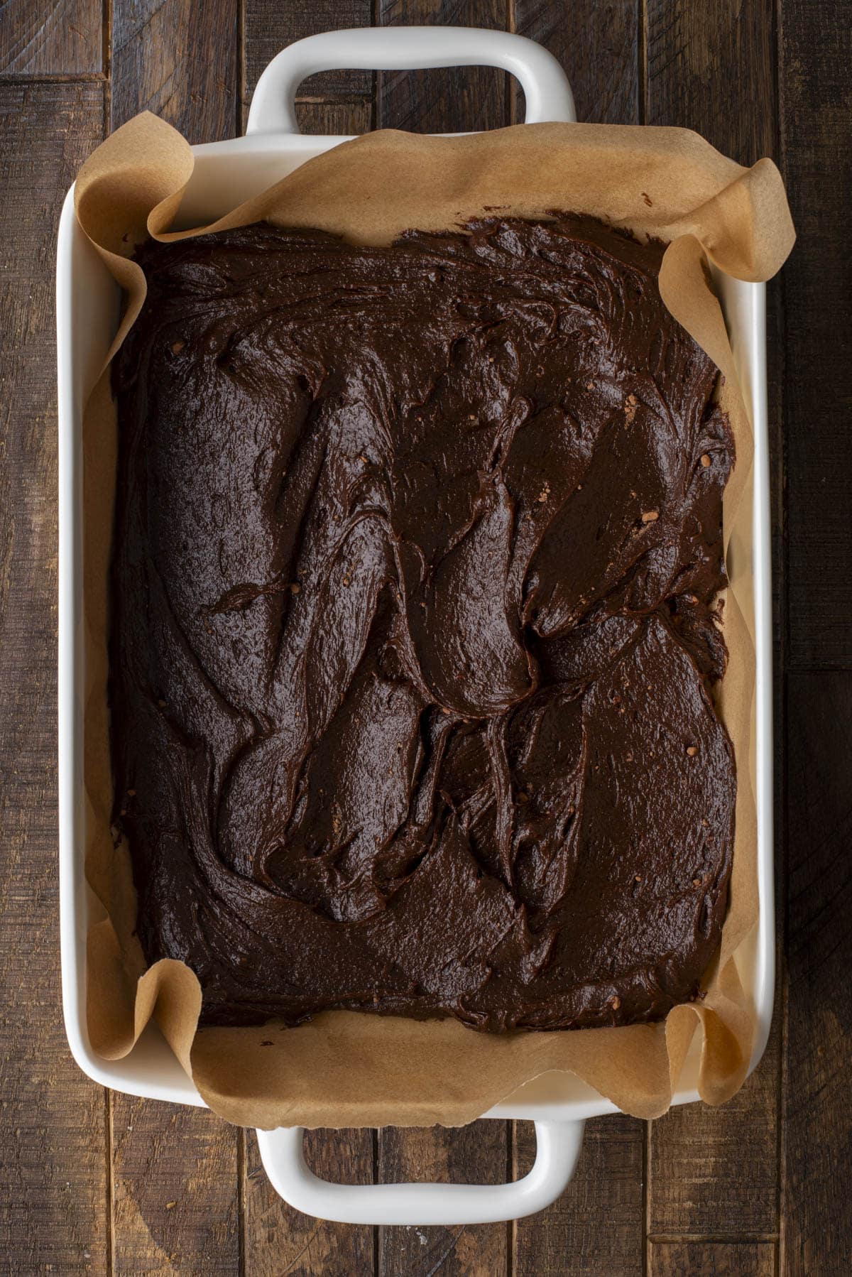 A white baking dish lined with brown parchment paper filled with unbaked chocolate brownie batter, set on a dark wooden surface.