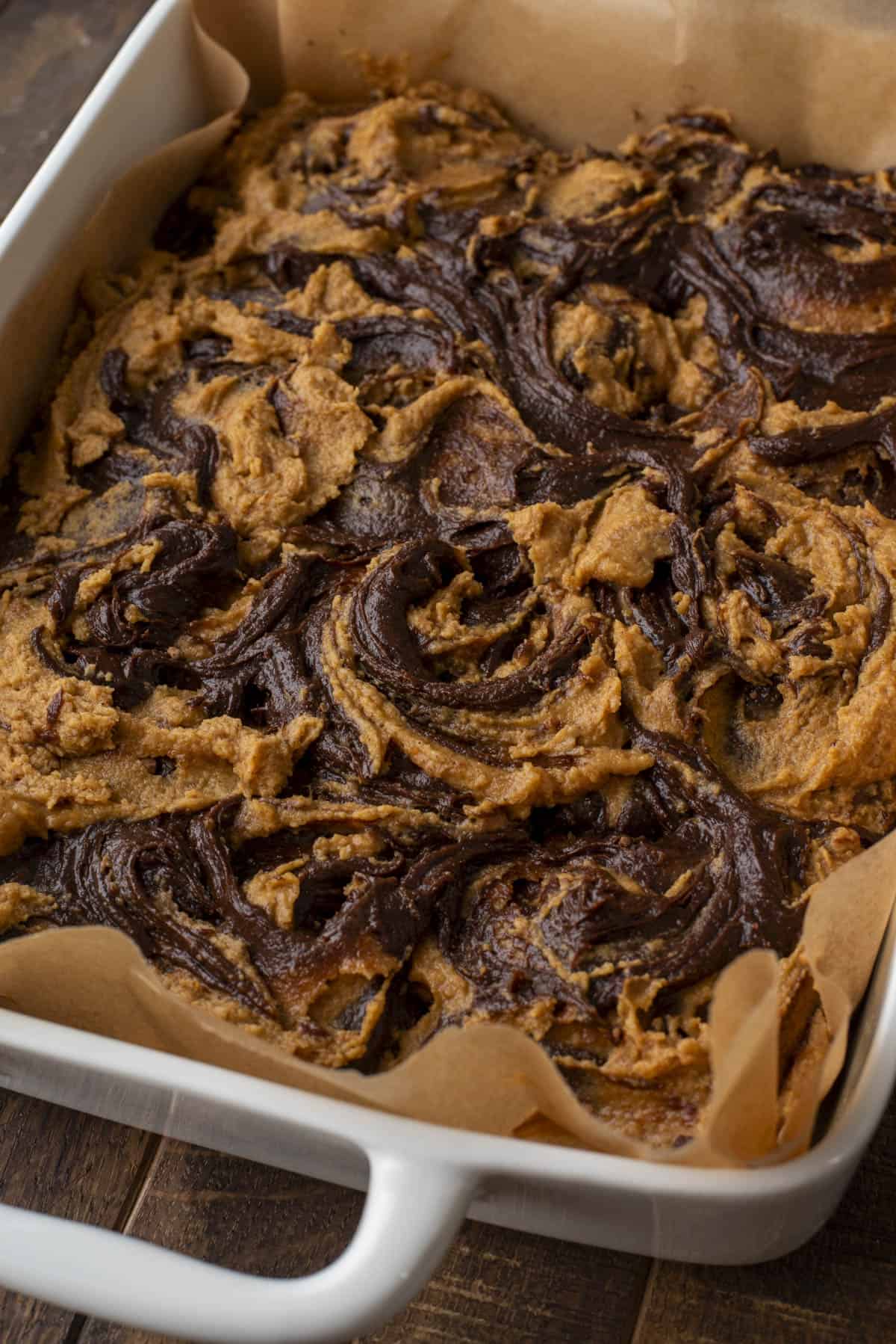 A close-up of a square baking dish lined with parchment paper, filled with a dessert featuring swirled layers of chocolate and peanut butter. The dessert is freshly baked and slightly golden at the edges.